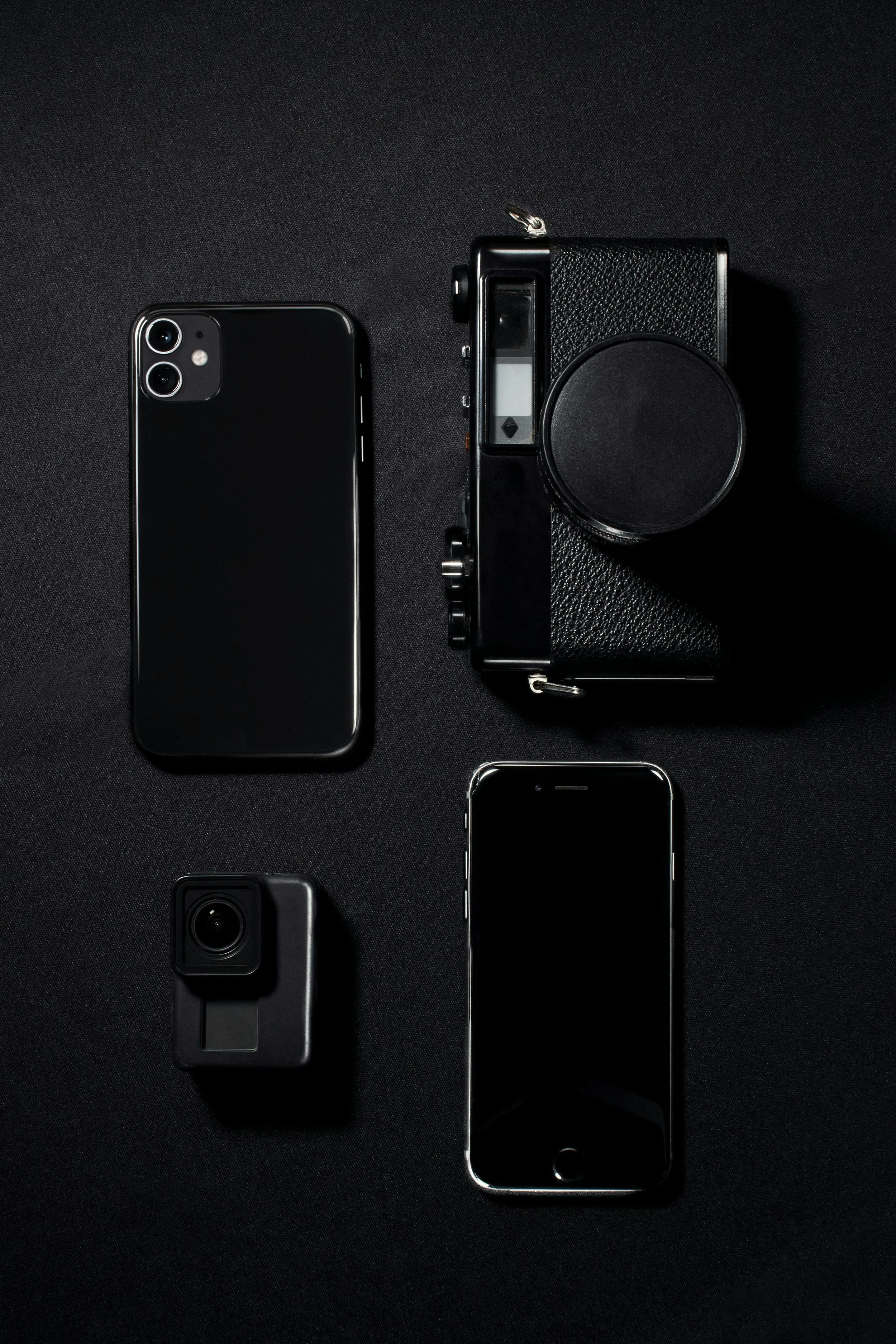 Collection of black electronic devices including a smartphone, a compact camera, a larger vintage camera, and a small action camera, all placed on a black surface.