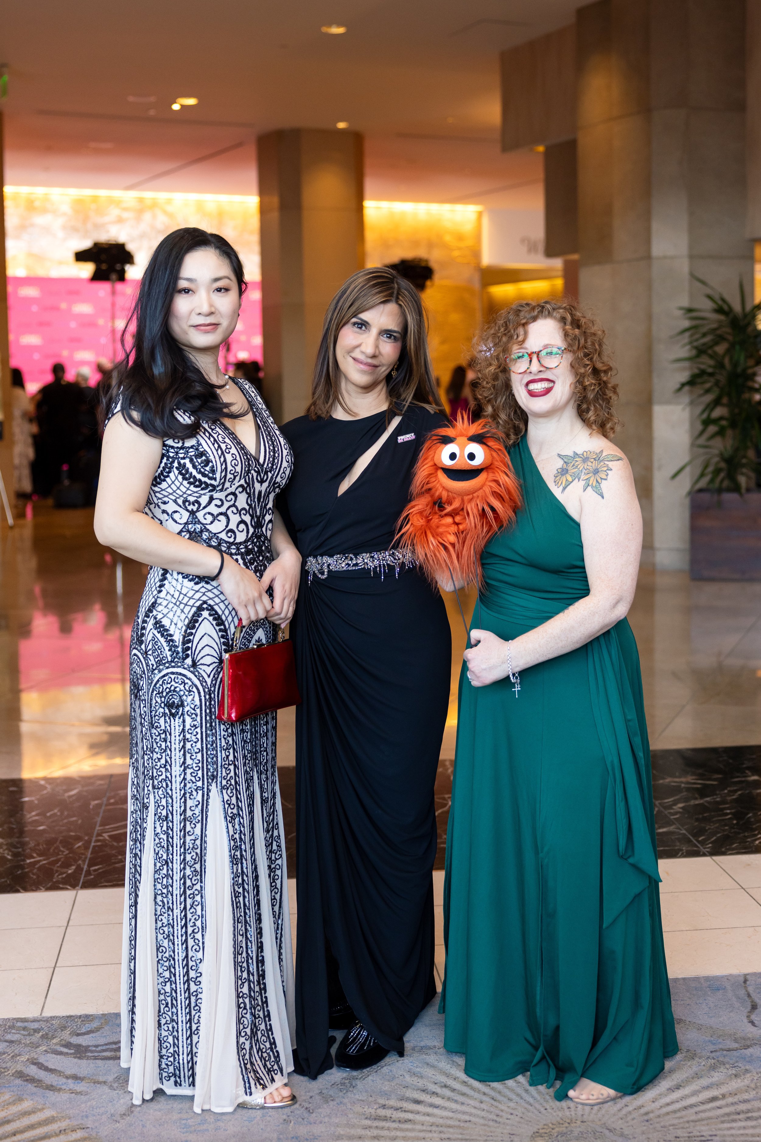 Three women standing together at an indoor event, dressed in formal evening wear. The woman on the left has long black hair and is holding a red clutch, wearing a white and navy patterned dress. The woman in the middle has shoulder-length brown hair, wearing a black dress, and has a sash with rhinestones. The woman on the right has curly red hair, glasses, and is wearing a green strapless dress, holding a puppet resembling a furry orange monster with a big smile.