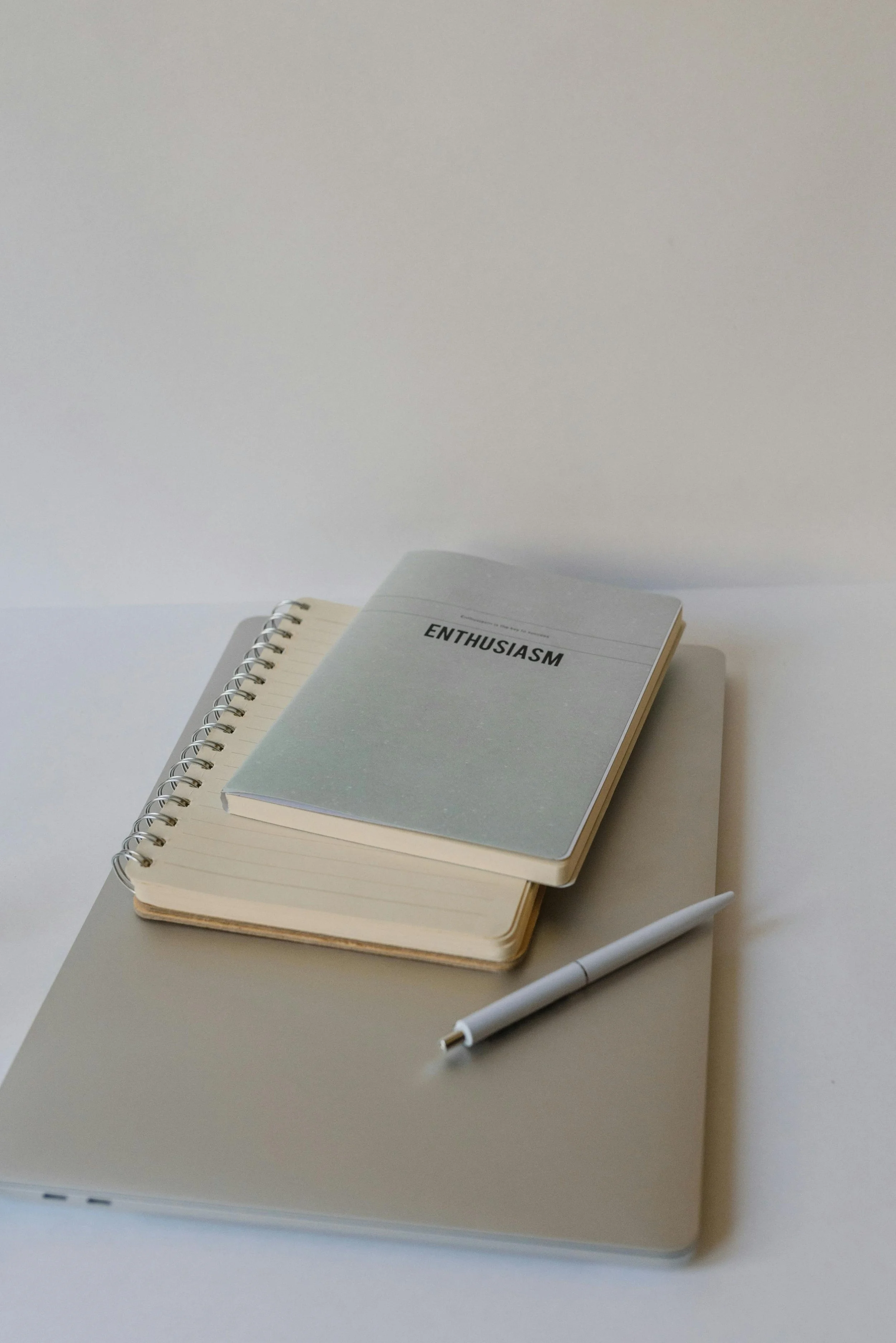 A stack of three notebooks and a white pen on a gray laptop, with the top notebook titled 'Enthusiasm' in black text on a gray cover.