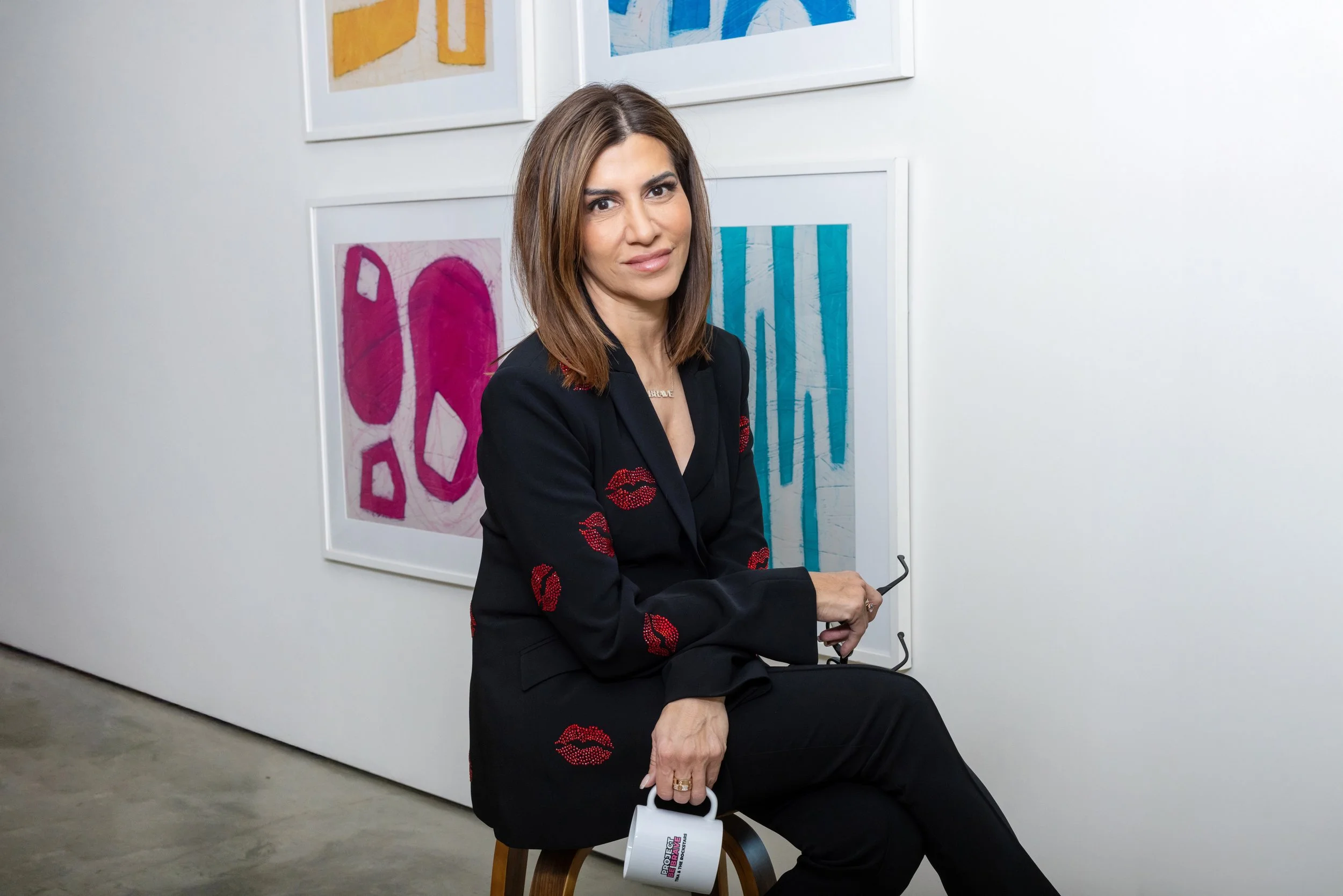 A woman with shoulder-length brown hair sitting on a chair in an art gallery, holding a white mug with a colorful logo, with colorful abstract artwork on the white wall behind her.