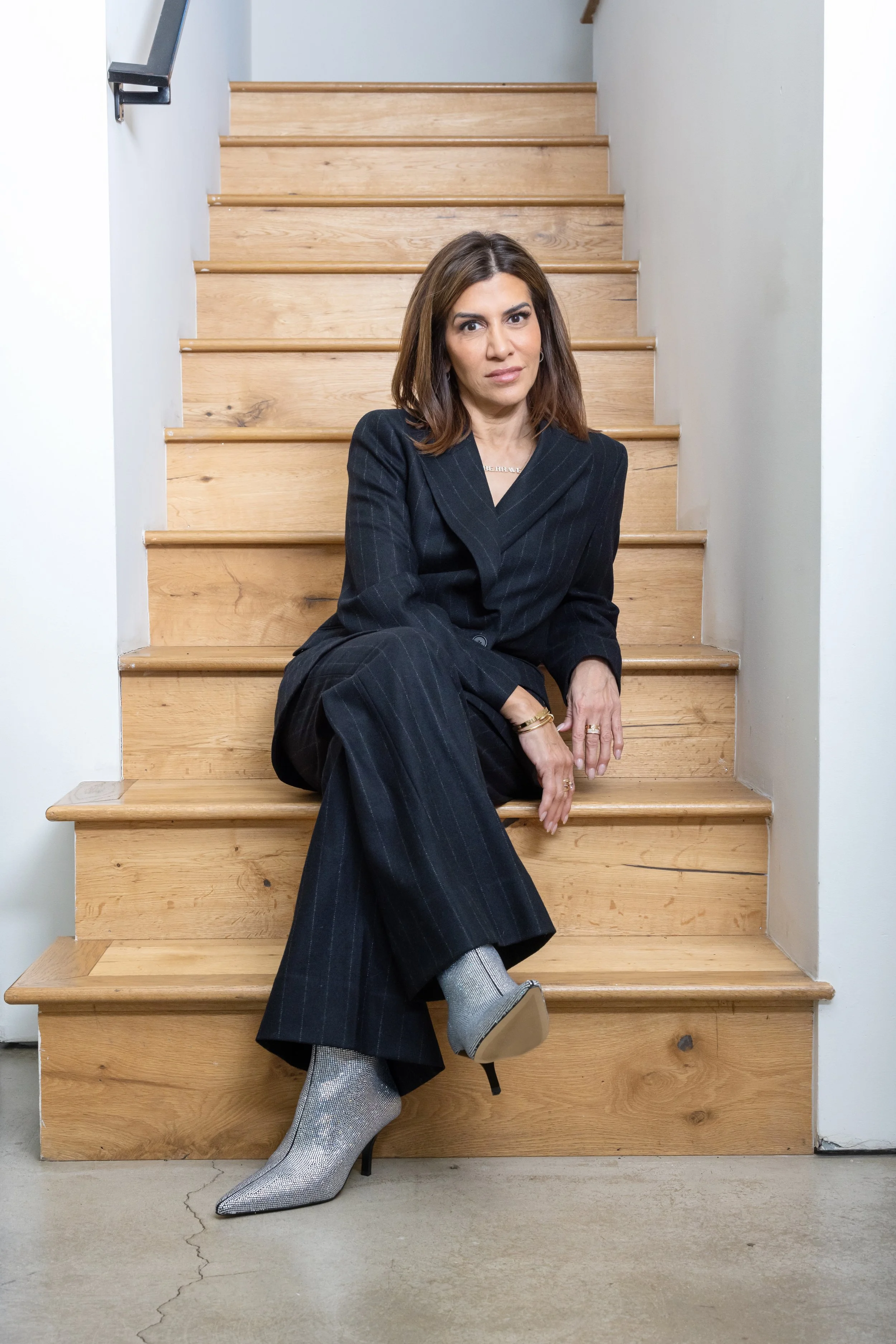 A woman in a dark pinstripe suit with silver high-heeled boots sitting on wooden stairs.