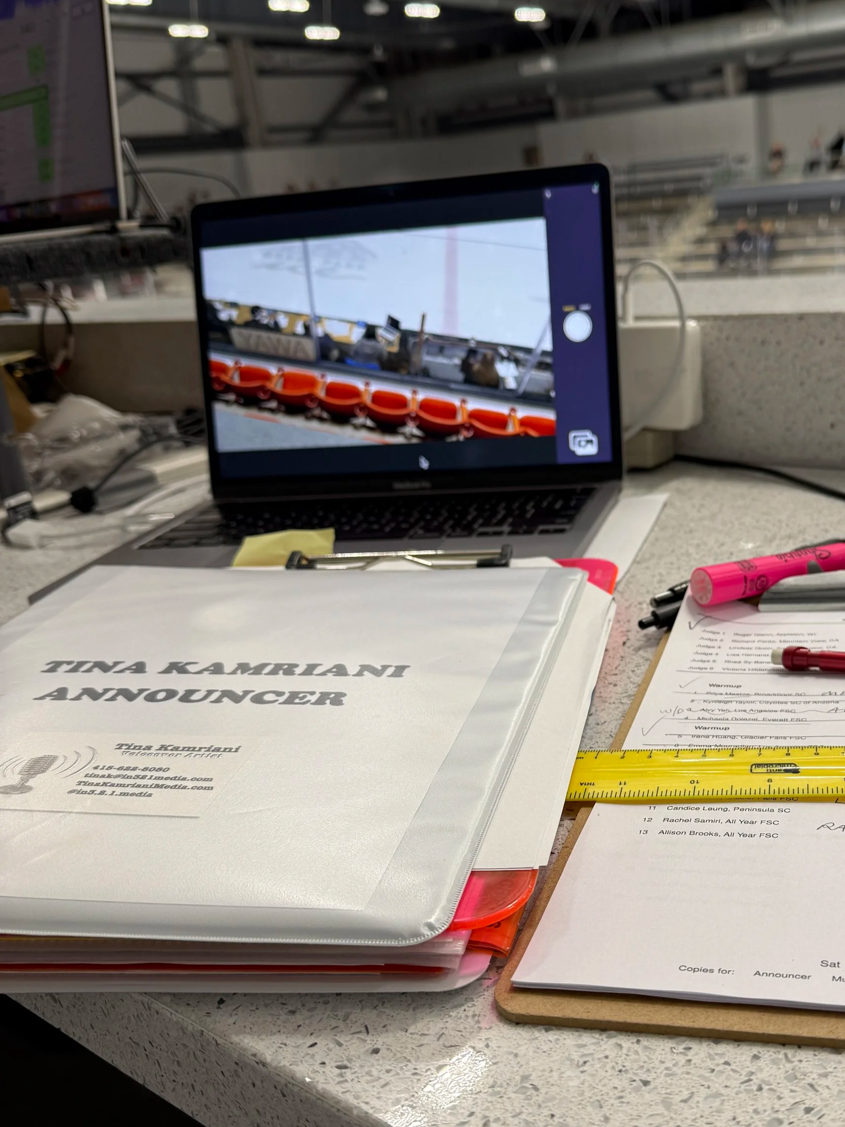 A desk with a folder labeled 'TINA KAMRIANT ANNOUNCER', a sheet of paper with notes, a yellow ruler, and a pink pen. A laptop is open, displaying a photo of a row of orange life preservers and some equipment against a sky background.