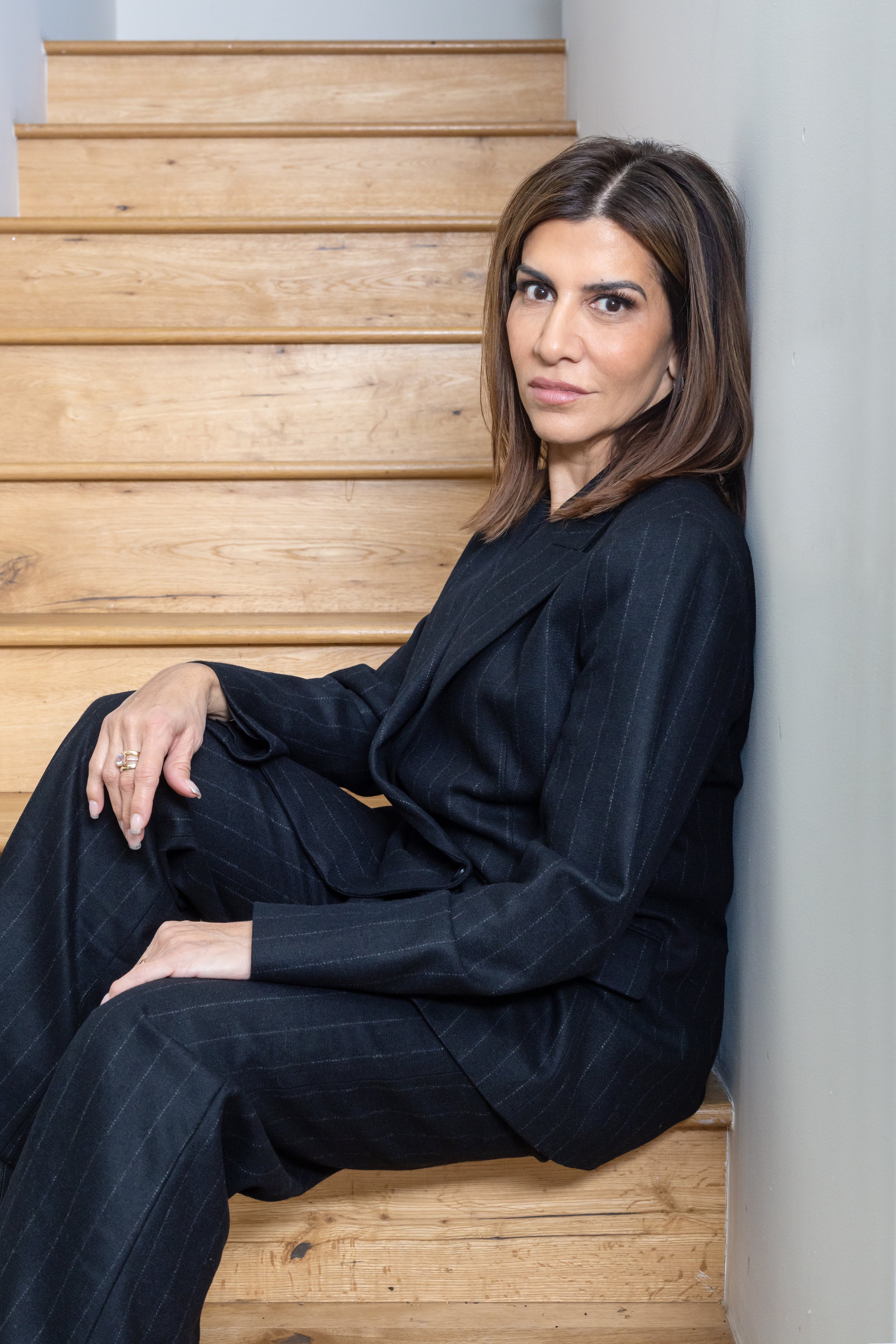 Woman with shoulder-length brown hair, wearing a dark pinstripe suit, sitting on wooden stairs and leaning against a white wall, looking at the camera.