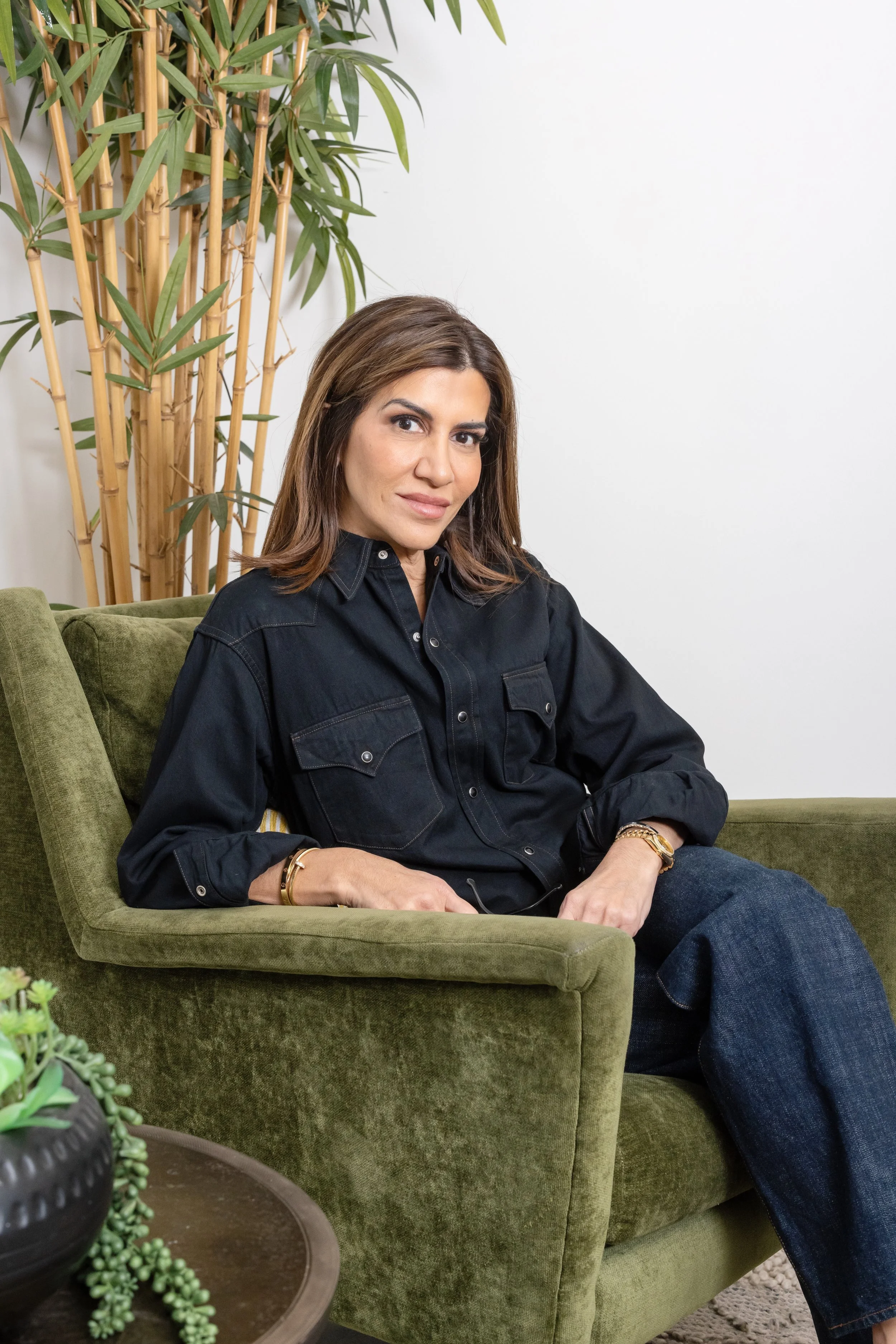 A woman with brown hair and a black shirt sitting on a green velvet sofa, with a tall houseplant behind her and a small table with a succulent plant in the foreground.