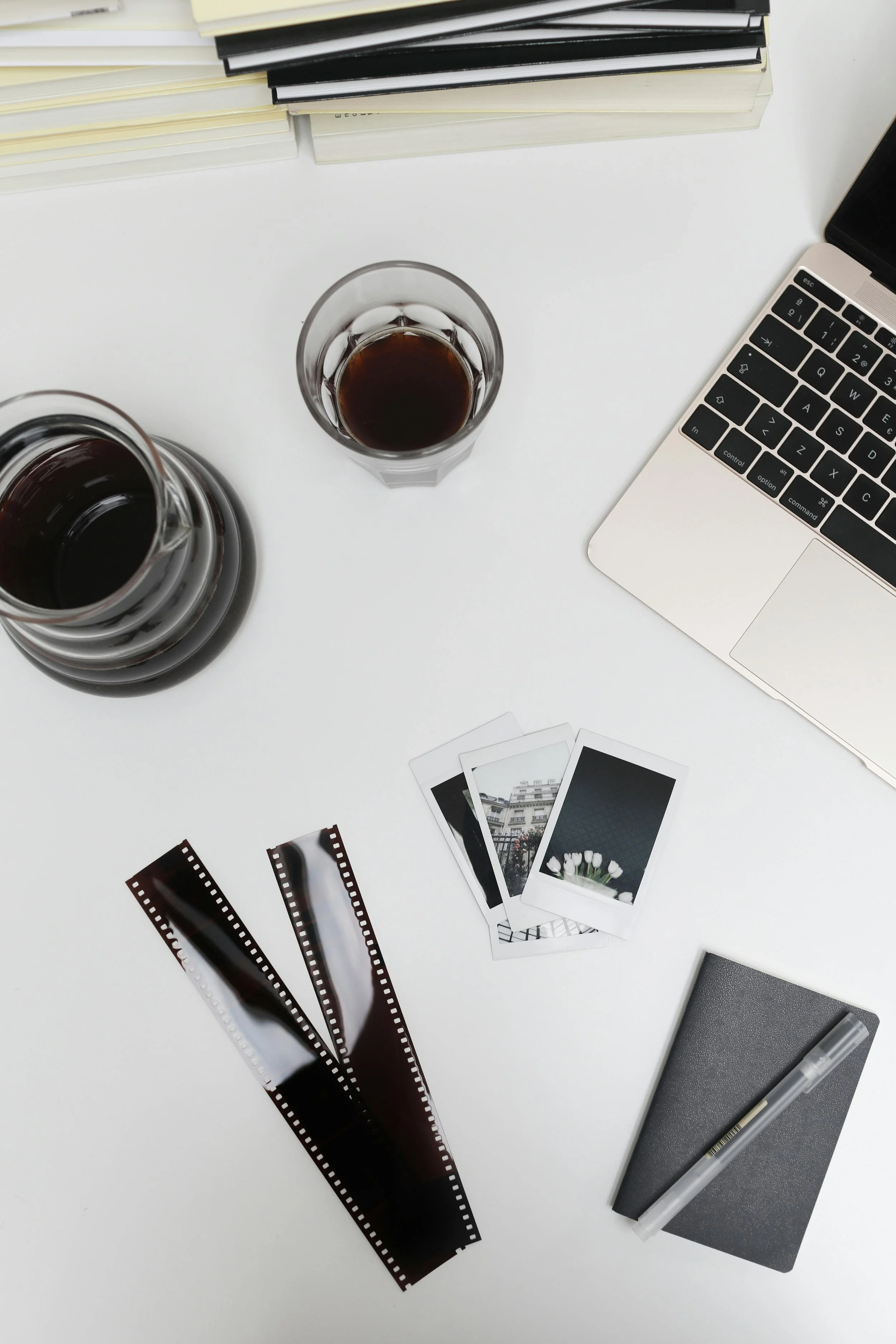 A workspace desk with a laptop, three film photos, two glasses of coffee, a gray notebook with a pen, a stack of books, a film strip, and some papers.