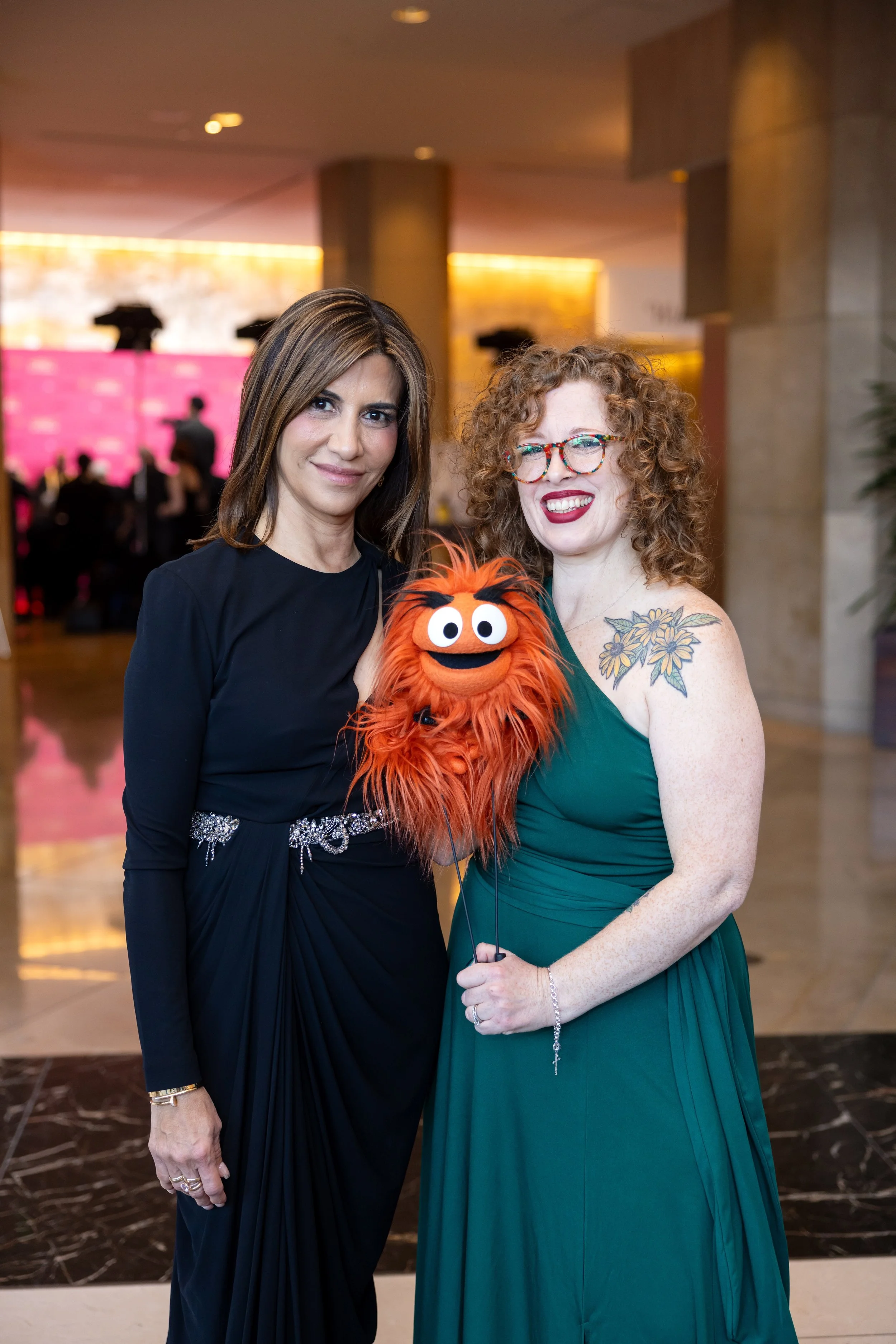 Two women in evening dresses holding a puppet with orange fur and wide eyes, smiling at an event with a pink background in the distance.