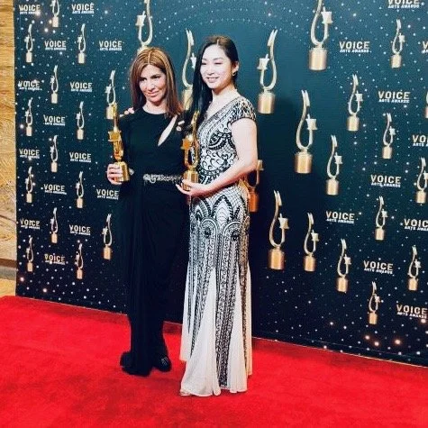 Two women standing on a red carpet at an awards event, holding trophies, with a backdrop featuring the word 'VOICE' and microphone graphics.