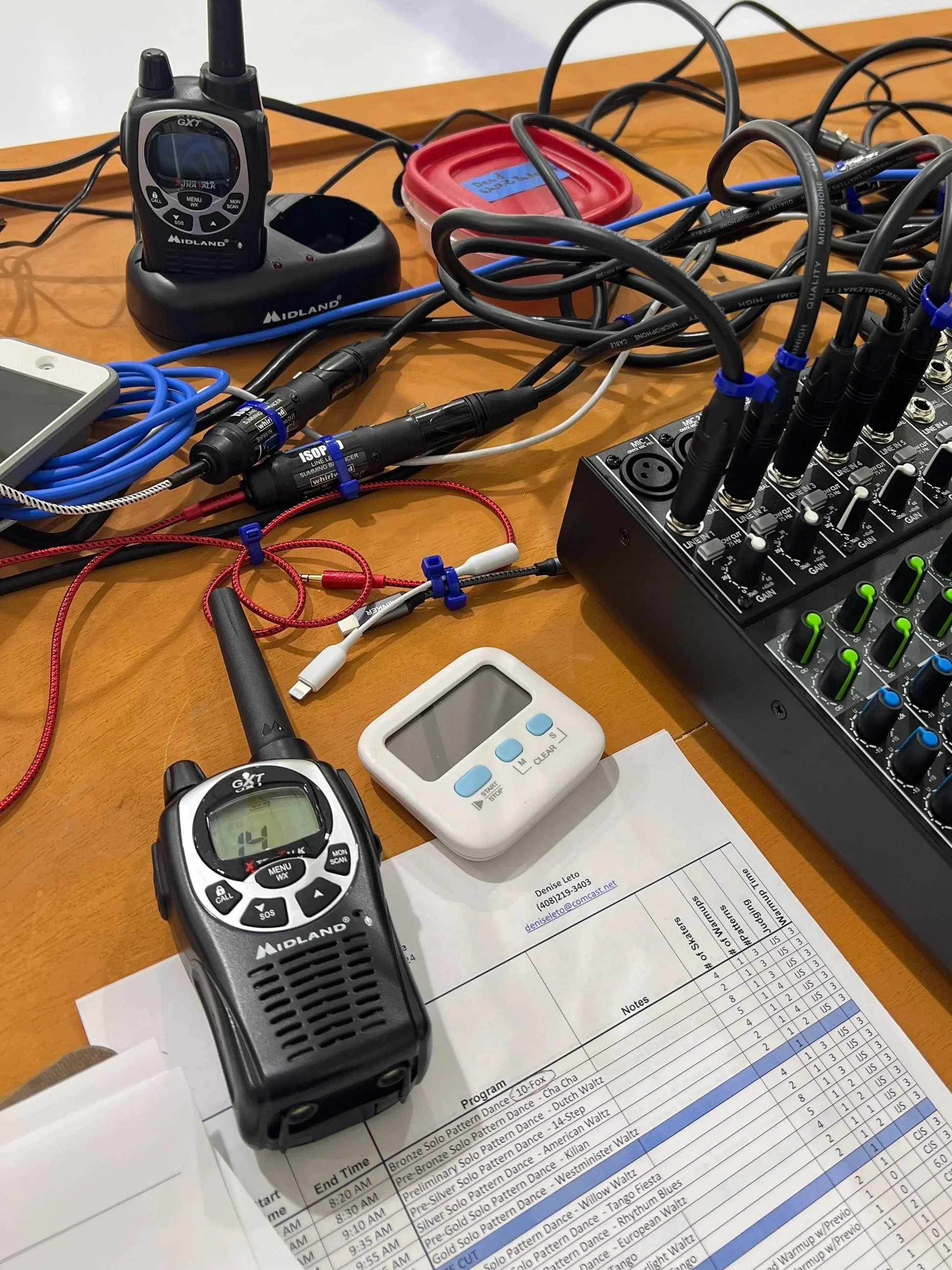 Musical equipment on a table including a wireless microphone, audio mixer, wired microphone, timing device, and various cables and accessories.