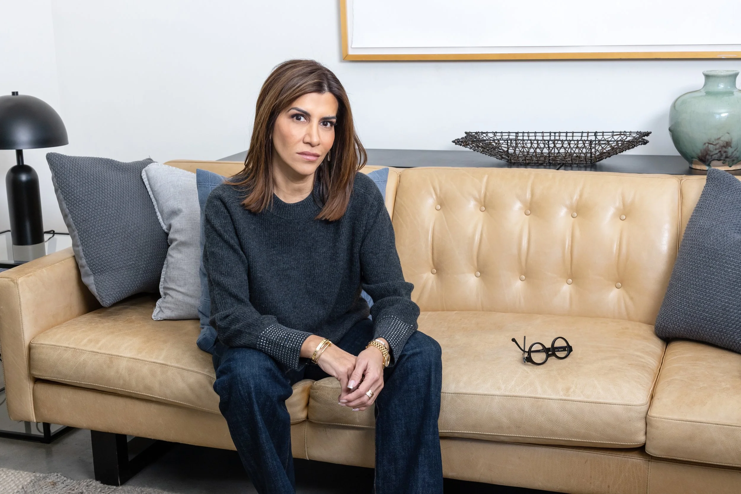 A woman sitting on a beige leather sofa in a modern living room with dark gray and light gray pillows, with a pair of black glasses on the sofa, and decorative items including a black wire tray and a green ceramic vase on a black shelf behind her.