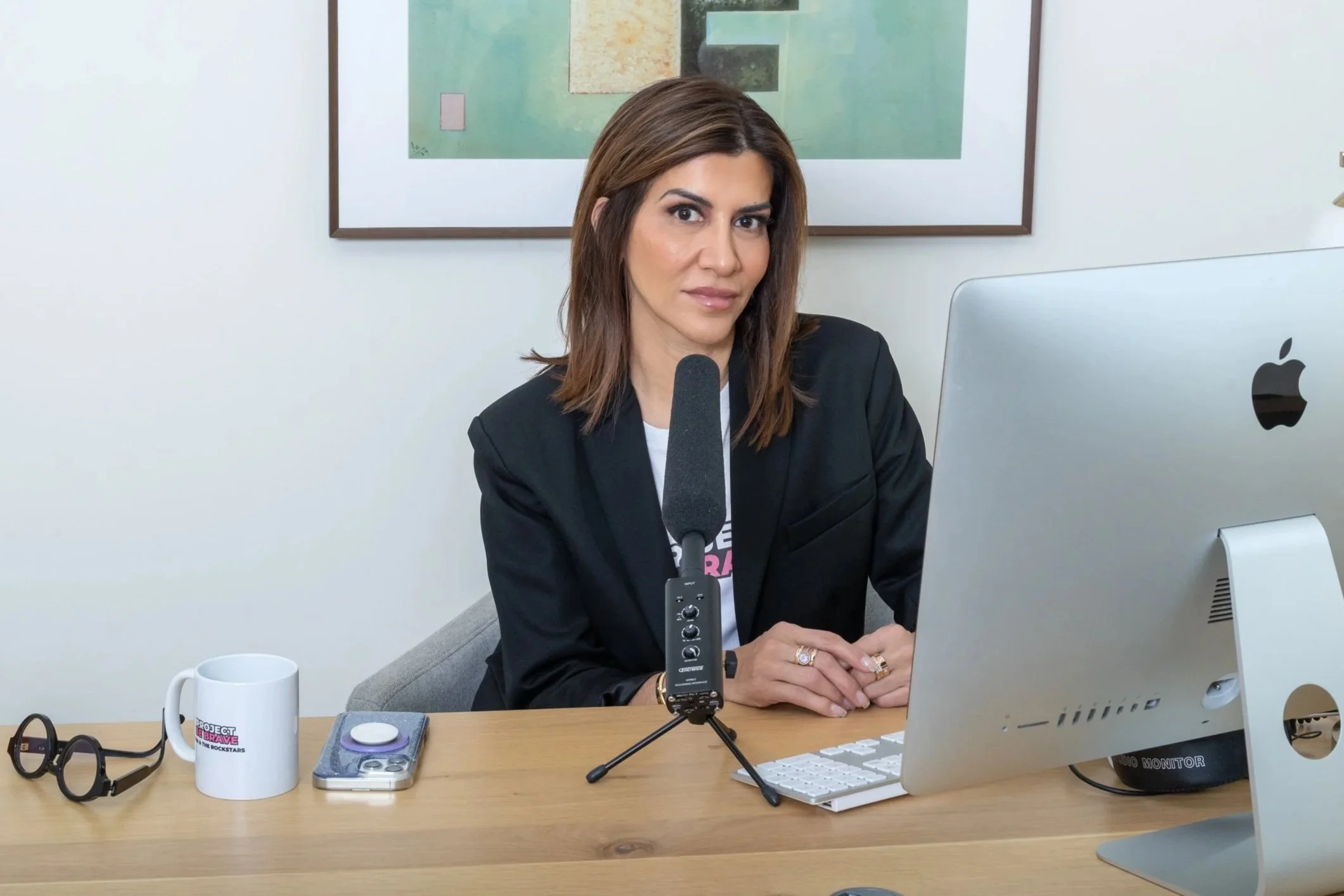 A woman with shoulder-length brown hair is sitting at a desk in front of a computer. She is wearing a black blazer and a white shirt. On the desk, there are a pair of glasses, a white coffee mug, a smartphone, and a small microphone on a stand. Behind her is a framed abstract art piece on the wall.