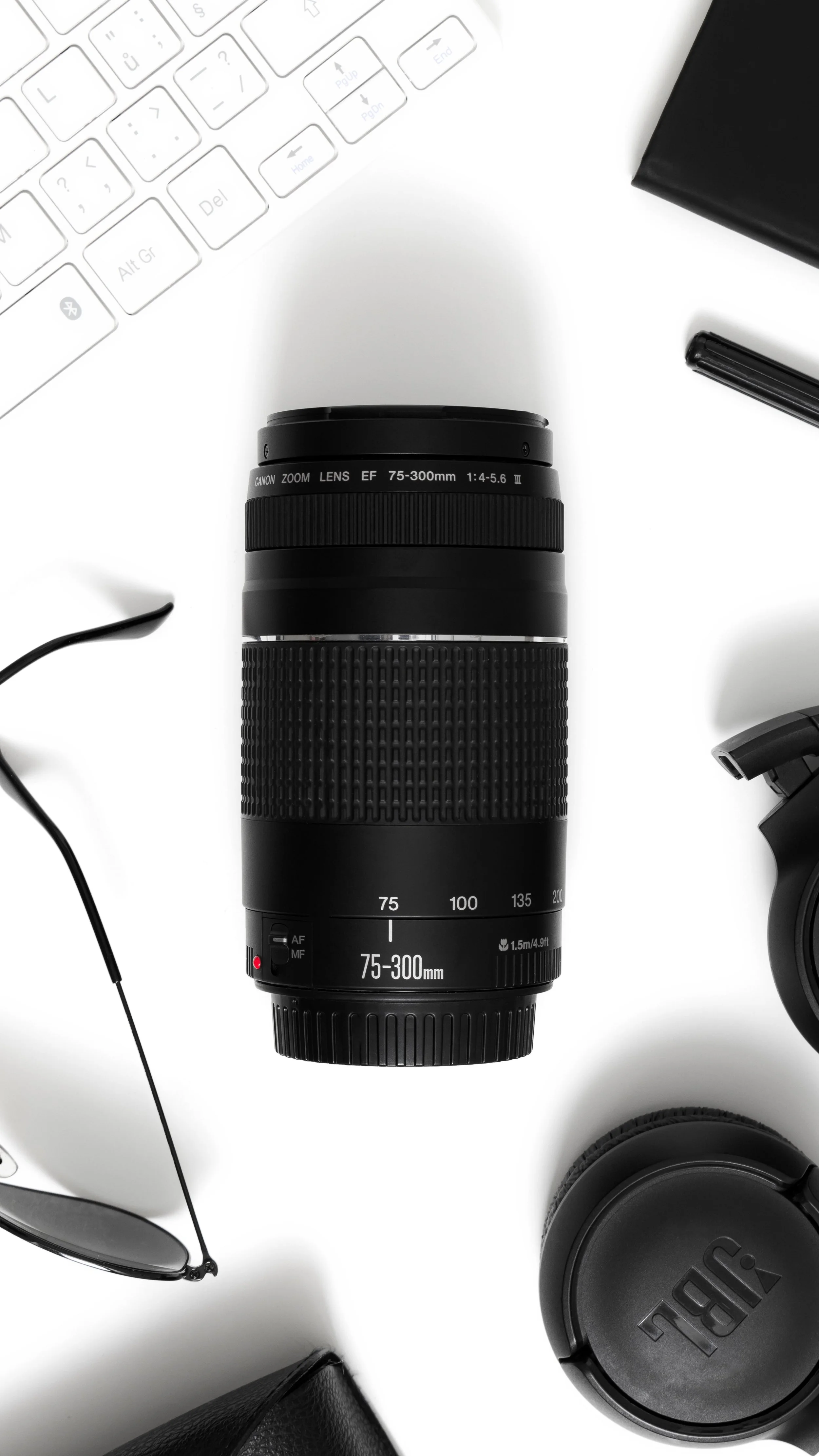 A Canon 75-300mm camera lens positioned on a white surface surrounded by a pair of sunglasses, a wireless keyboard, a black notebook, a black pen, a speaker, and a camera viewfinder.