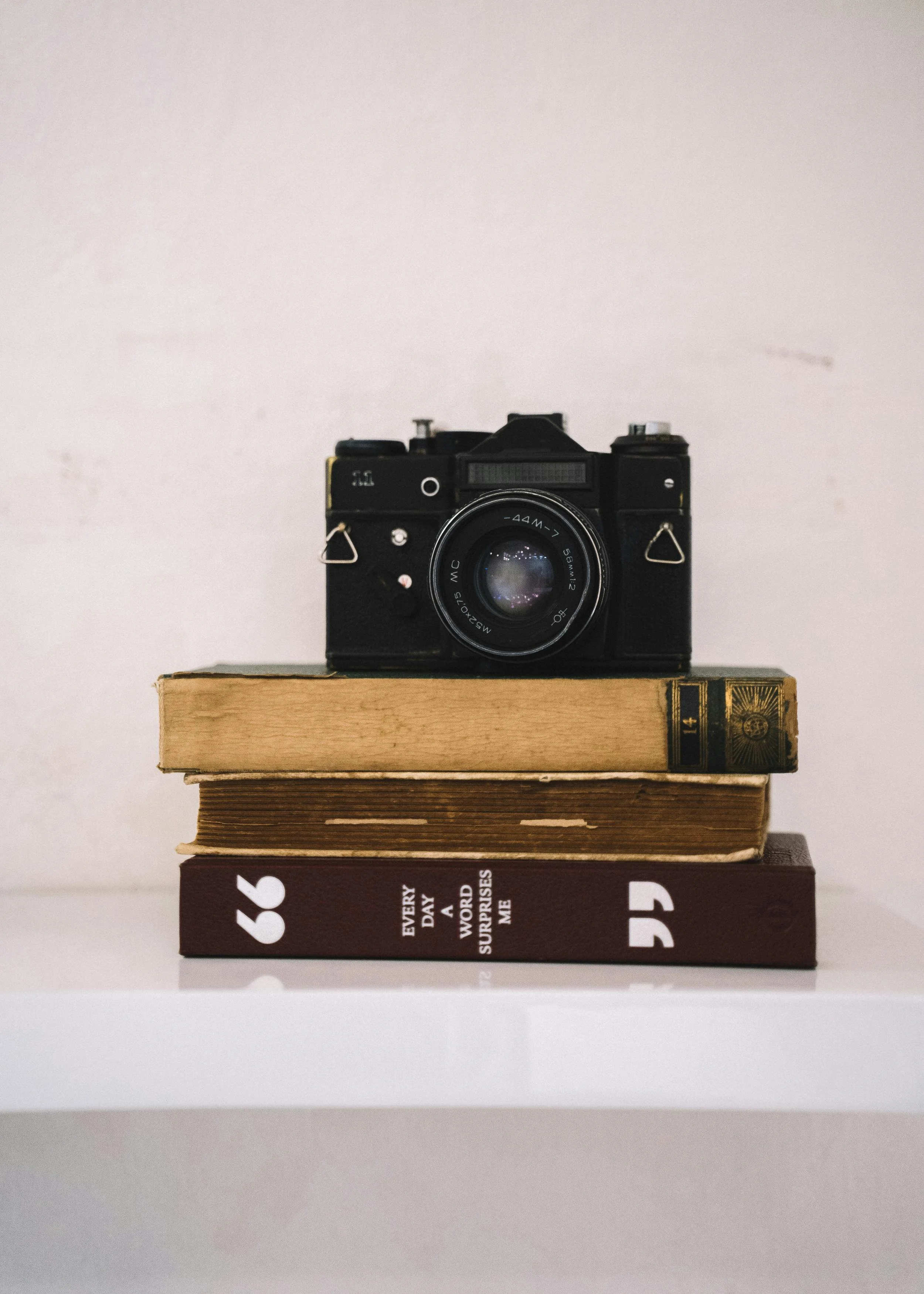 A vintage black camera placed on top of two stacked books on a white shelf against a plain background.
