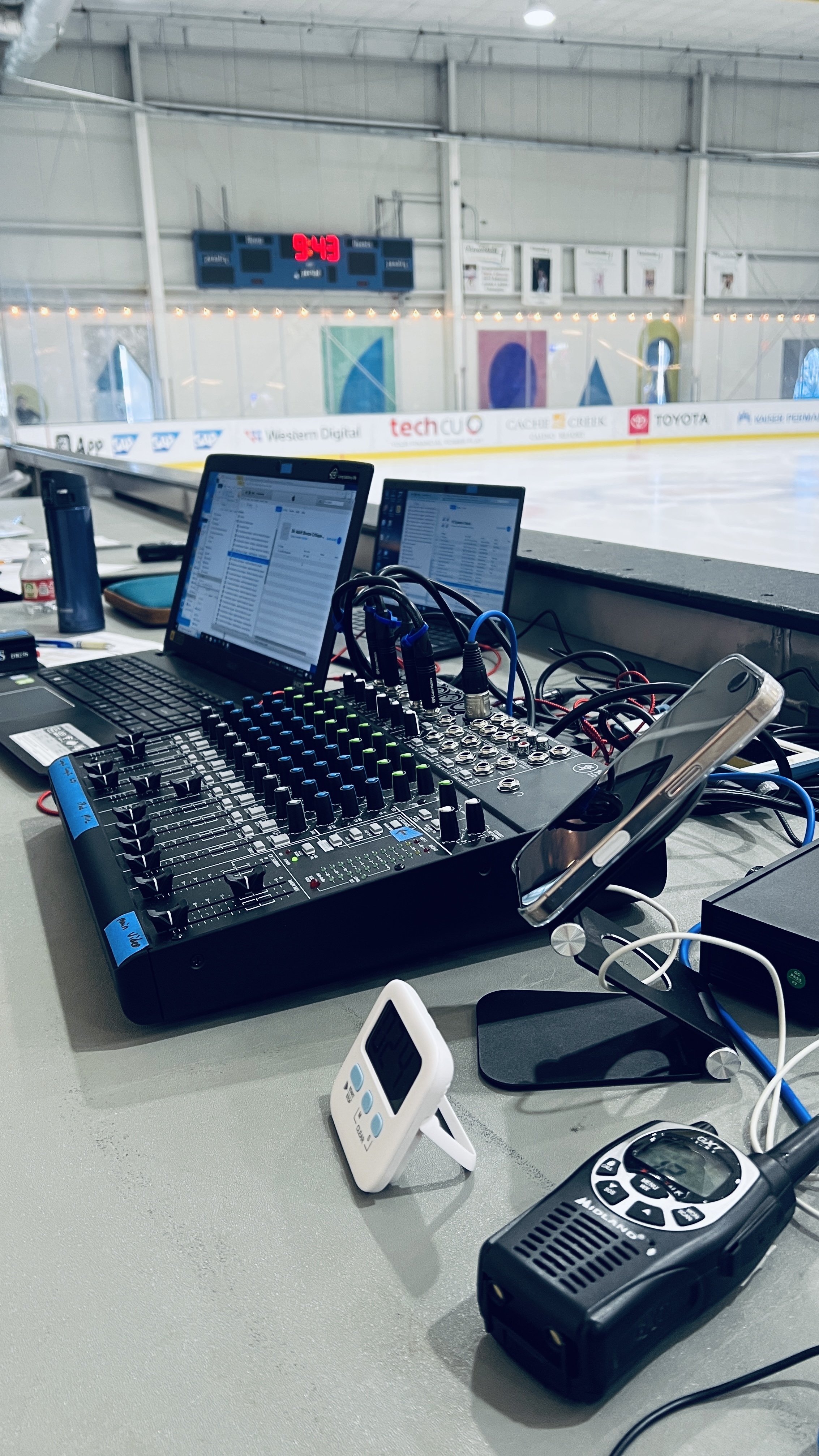 A scoreboard and equipment on a table at an indoor ice hockey rink, with digital screens, a mixer, radio, and timers.