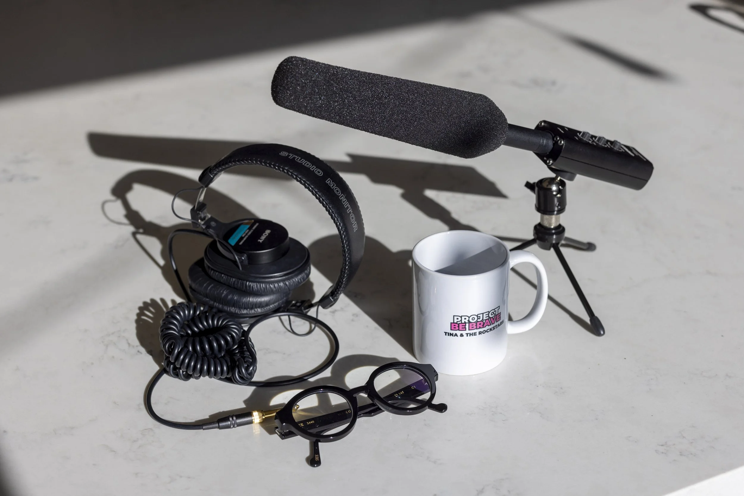 A white coffee mug with the text 'Project Be Brave Tina & the Rockstars' printed on it, a pair of black over-ear Sony headphones, a pair of black round glasses, a professional microphone with a foam windscreen on a small tripod, and a pair of glasses are arranged on a white surface with natural sunlight creating shadows.