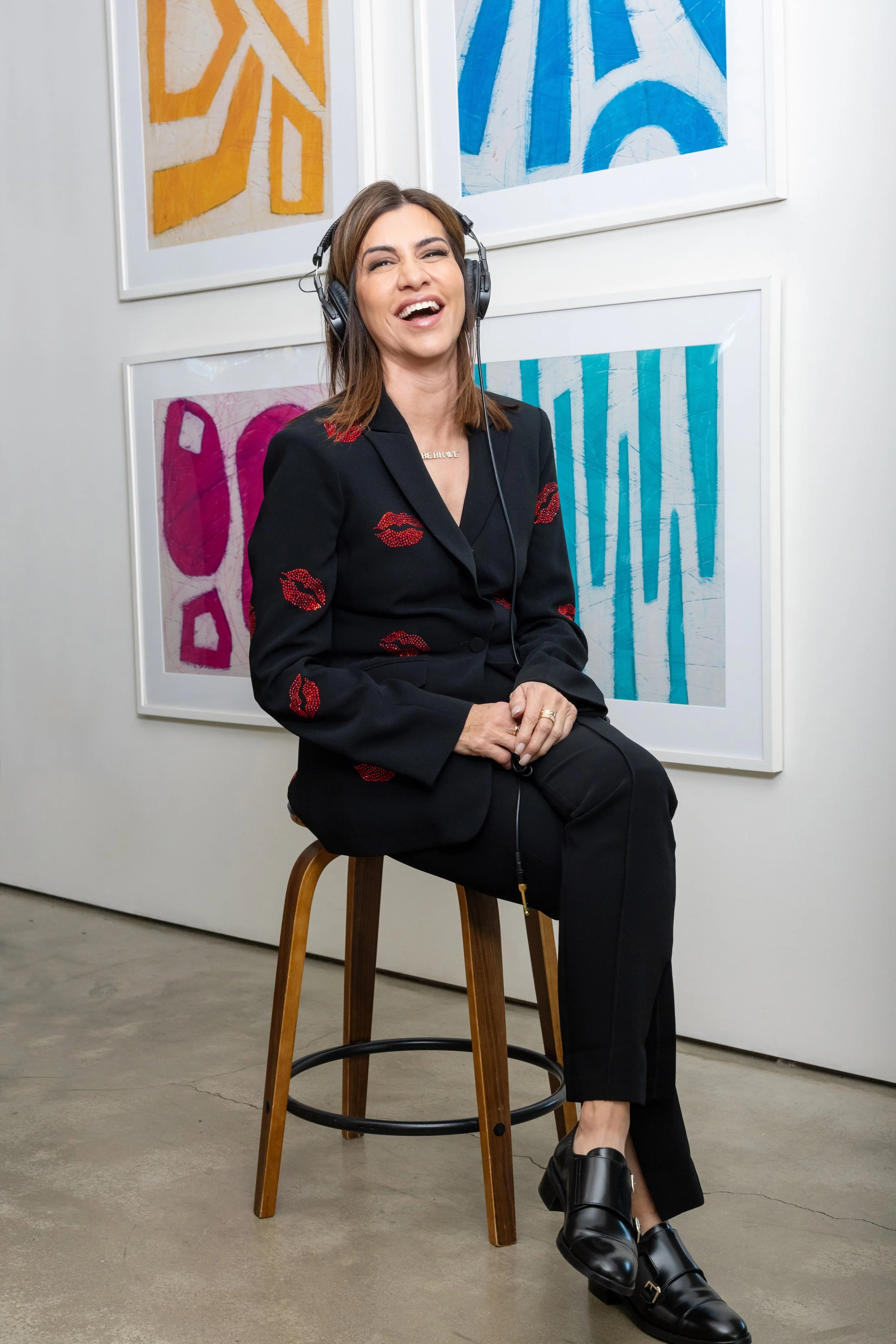 A woman wearing a black blazer with red lips patterns, sitting on a wooden stool, laughing with headphones on, in front of abstract artwork on the wall.