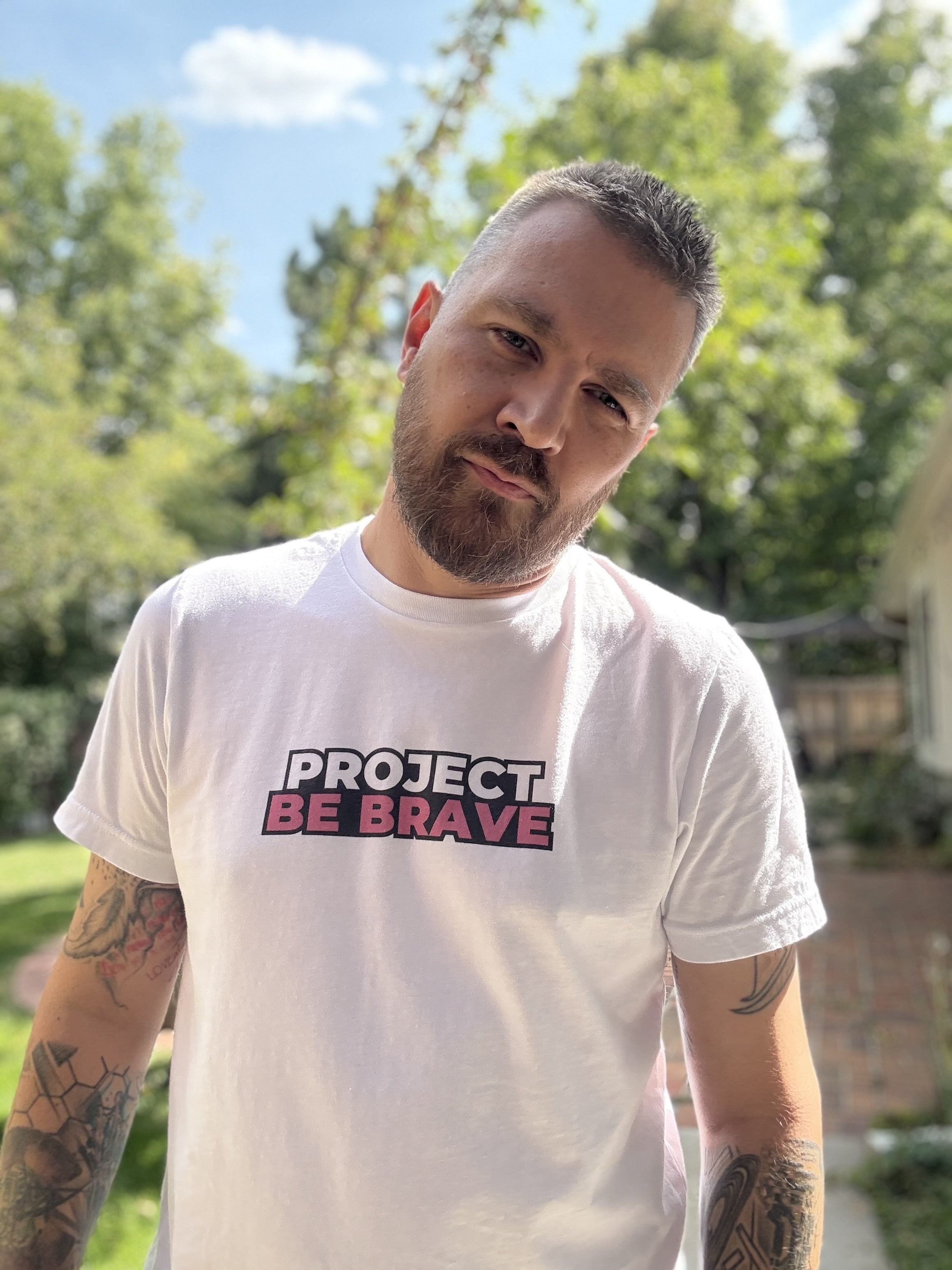 A man with short hair and a beard wearing a white T-shirt with the words 'PROJECT BE BRAVE' in black and pink, standing outdoors with trees and a house in the background.