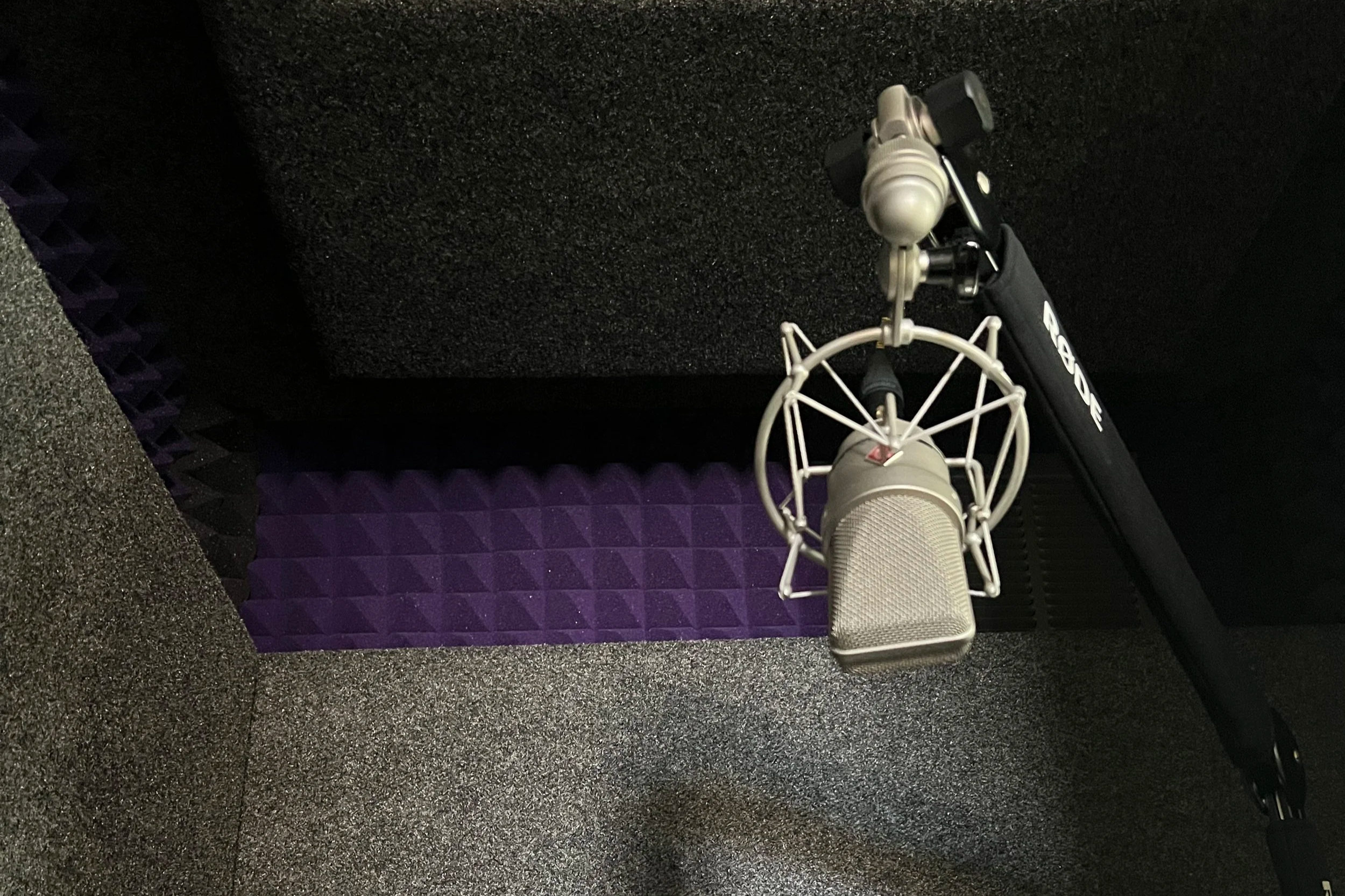 Close-up of a silver microphone with a shock mount, set up in a recording booth with foam soundproofing panels on the wall and floor.