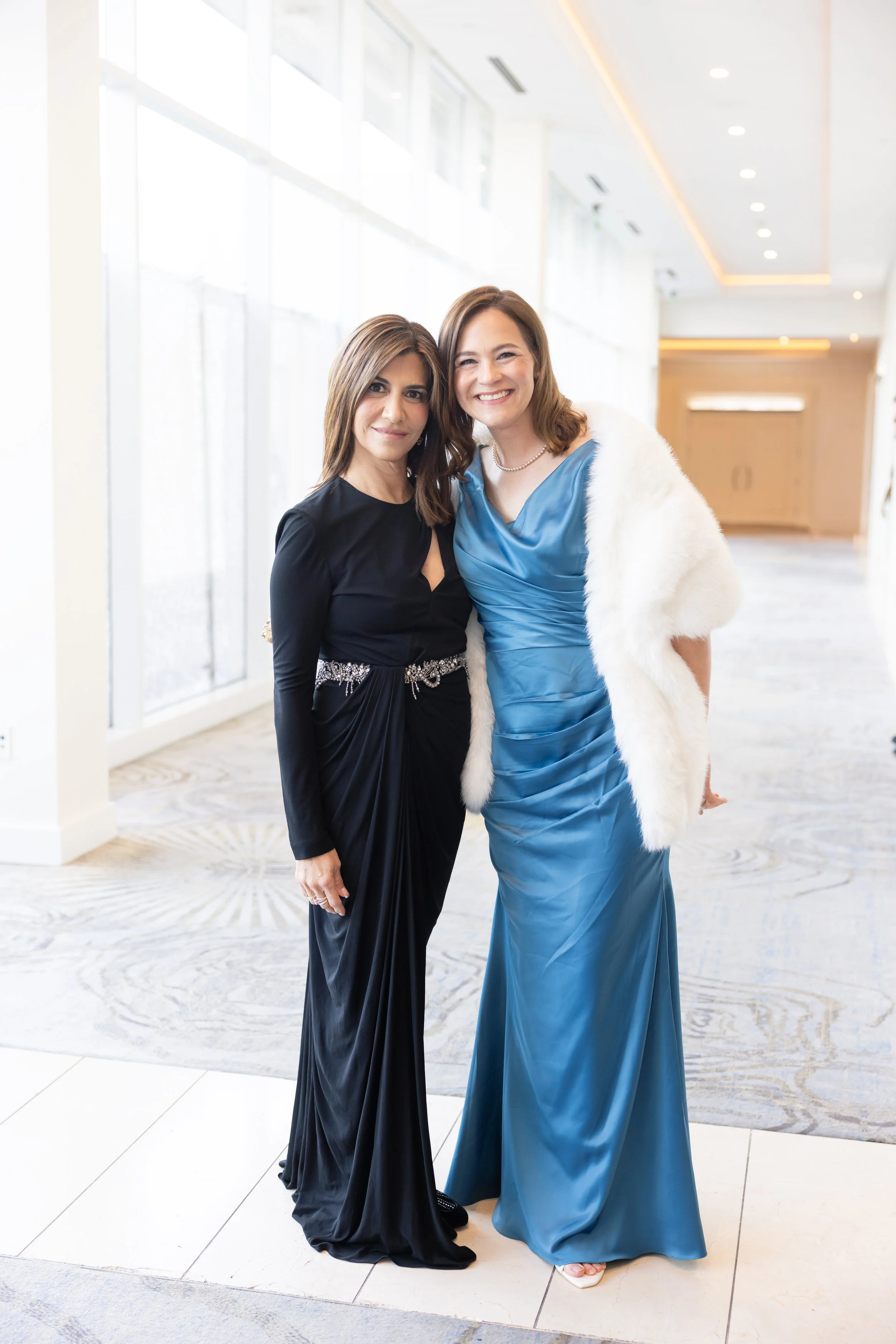 Two women in formal dresses standing close together and smiling at the camera in a brightly lit indoor space.