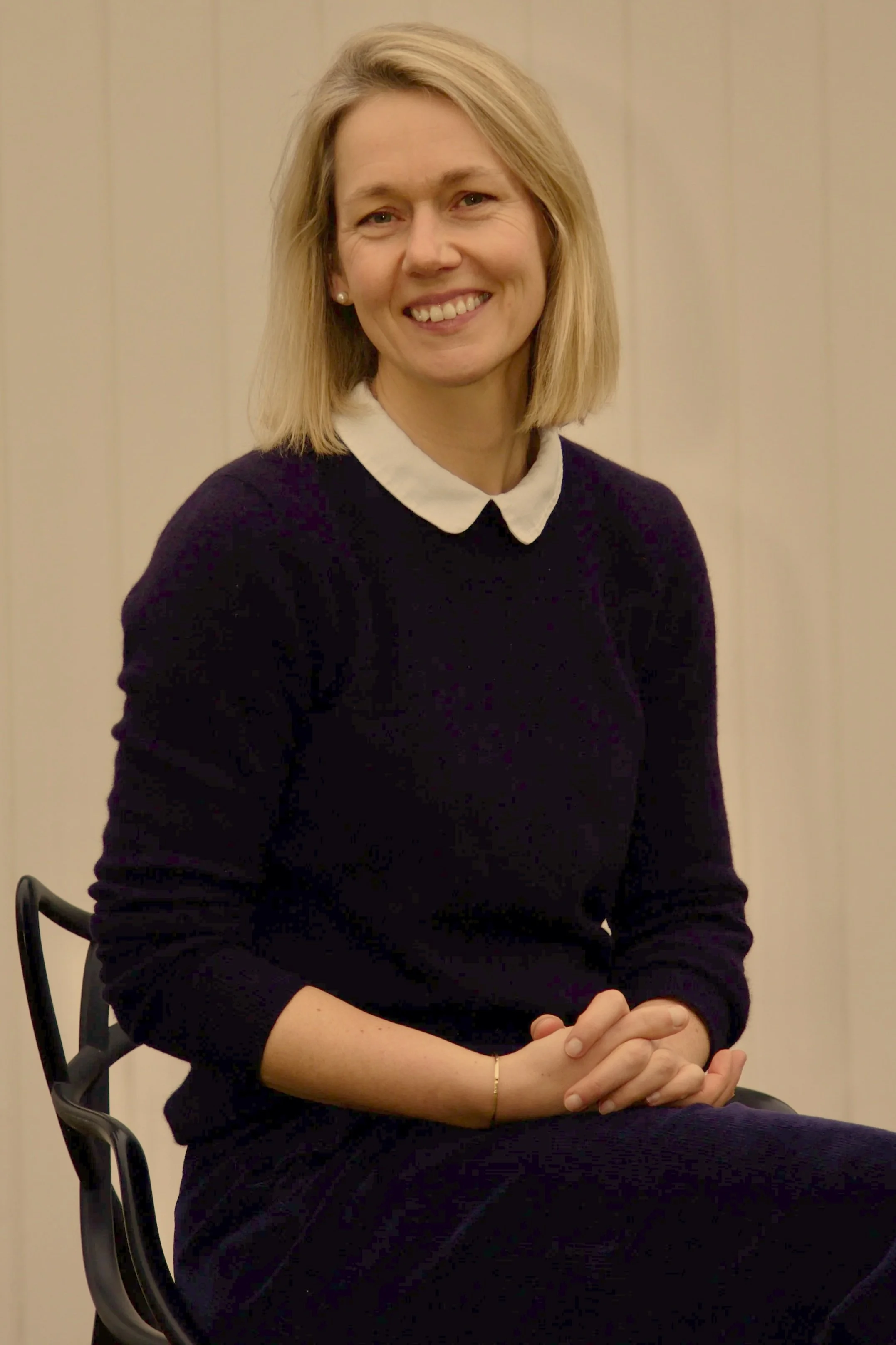 A woman with blonde hair, wearing a black sweater and a white collared shirt, sitting on a chair with hands clasped in her lap, smiling at the camera.