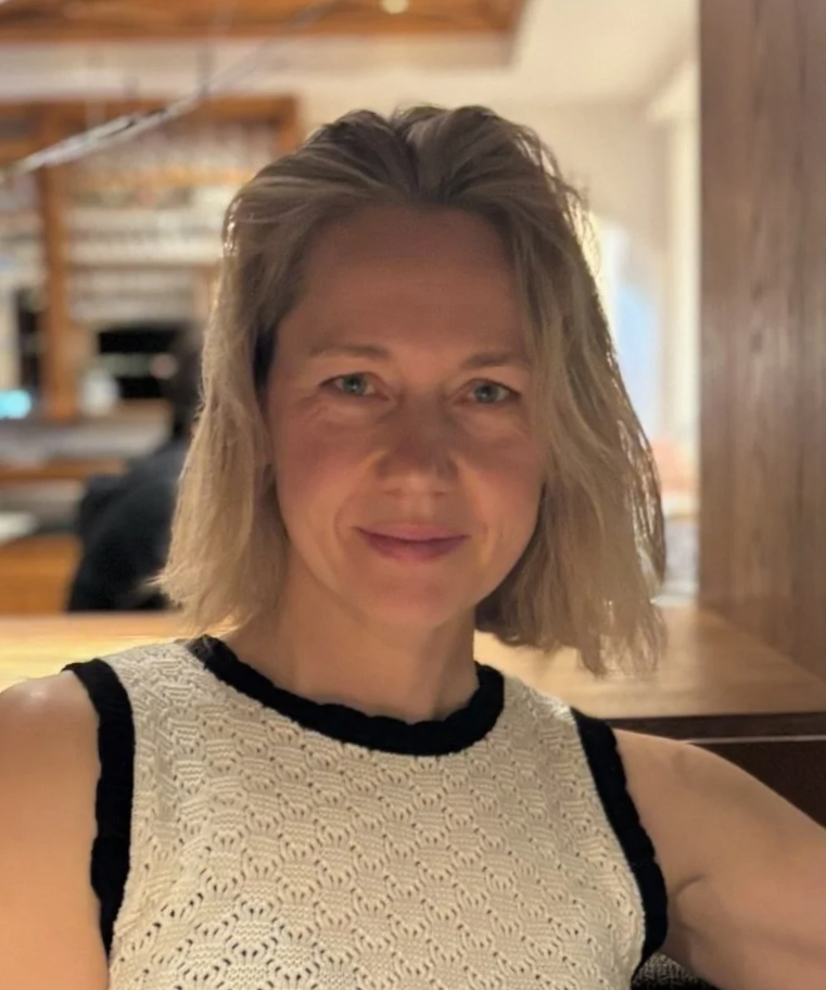 A woman with short blonde hair, wearing a sleeveless white knitted top with black trim, sitting indoors with a blurred wooden interior background.