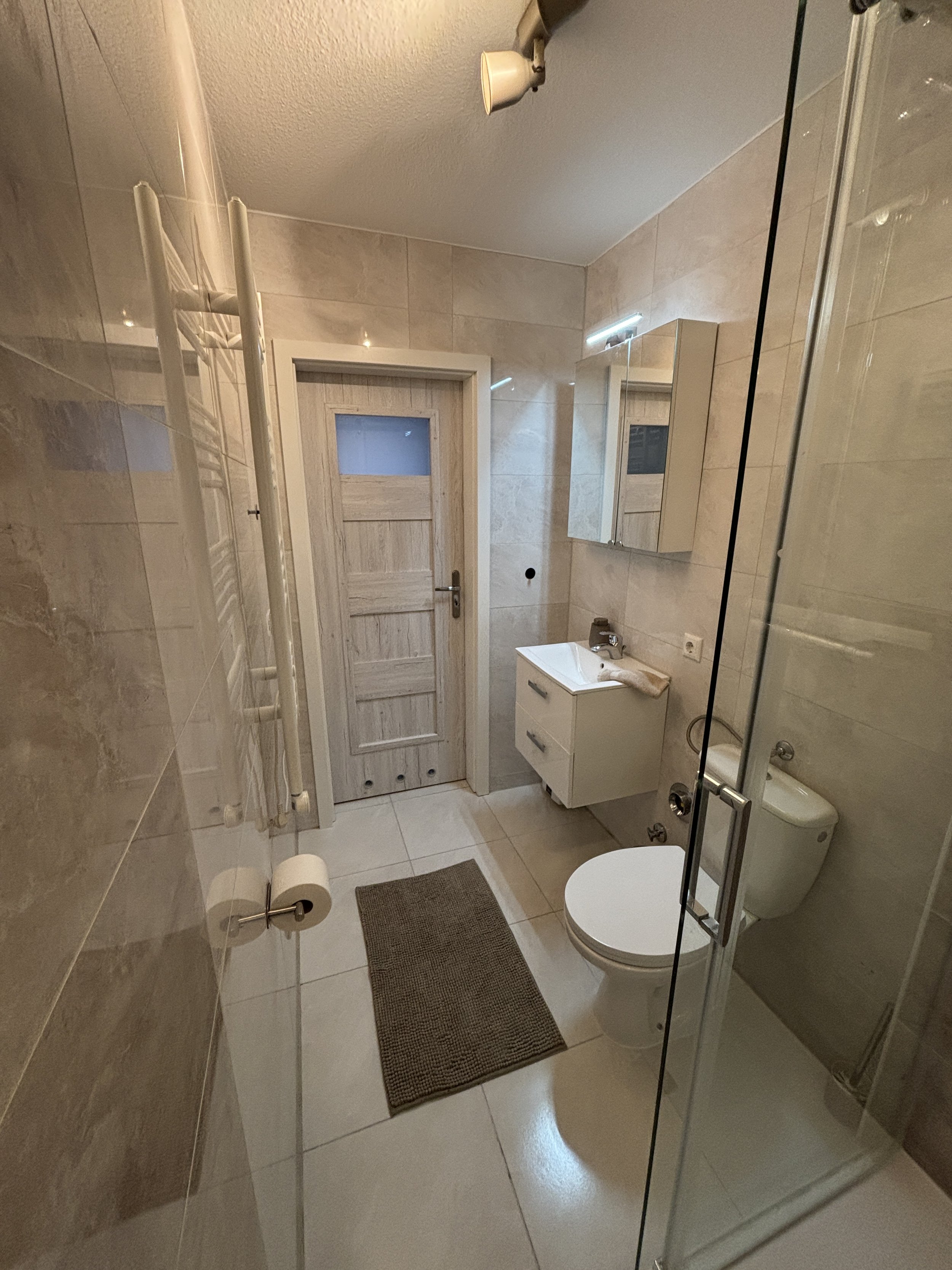 Ein kleines Badezimmer mit Toilette, Waschbecken, Spiegel, Handtuch, Badewanne, Beleuchtung, Tür und Fliesen.