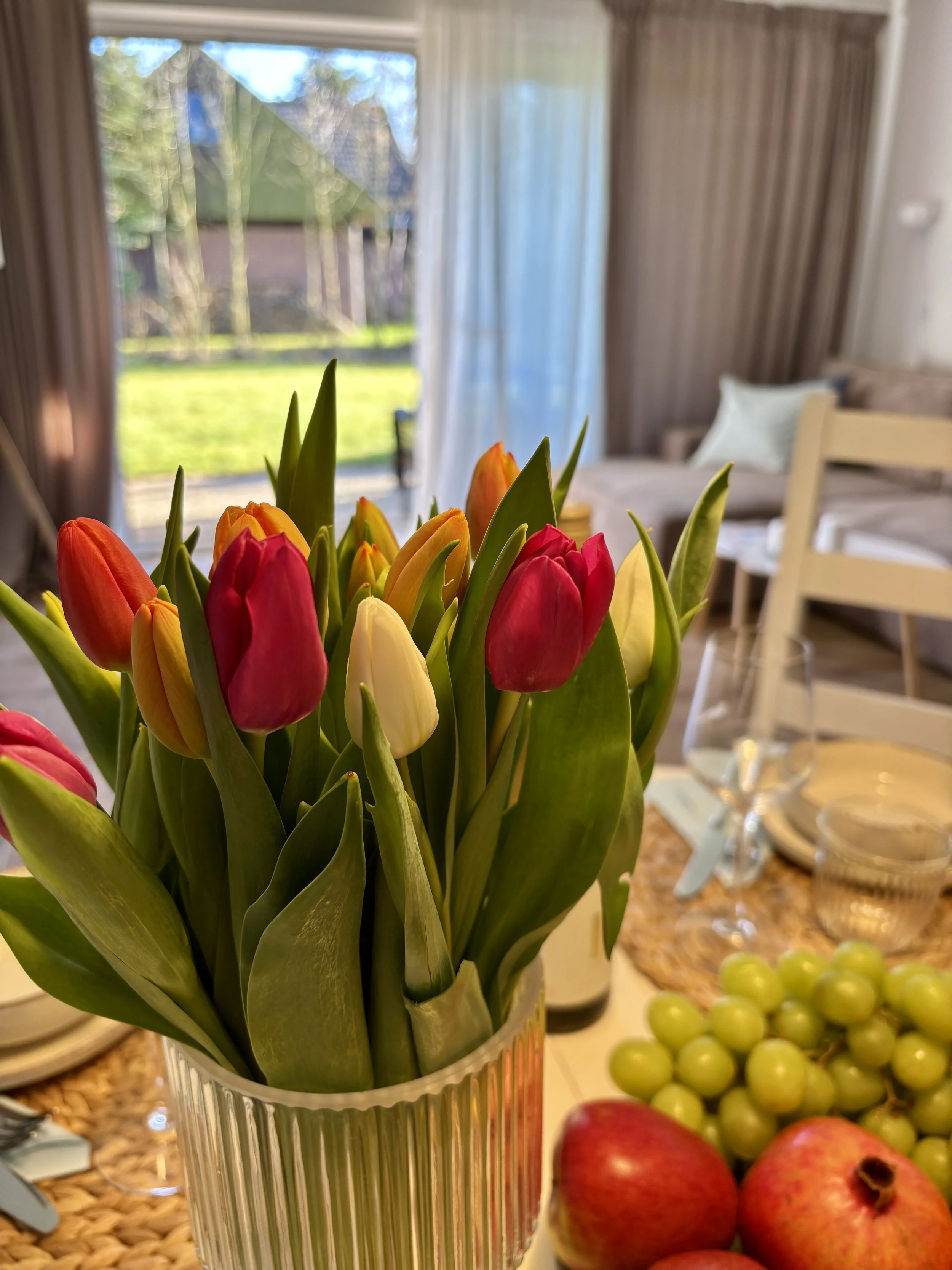 Bunter Tulpenstrauß in einer Vase auf einem gedeckten Tisch, mit Früchten und Geschirr, im Wohnzimmer mit großer Fensterfront und Blick ins Freie.
