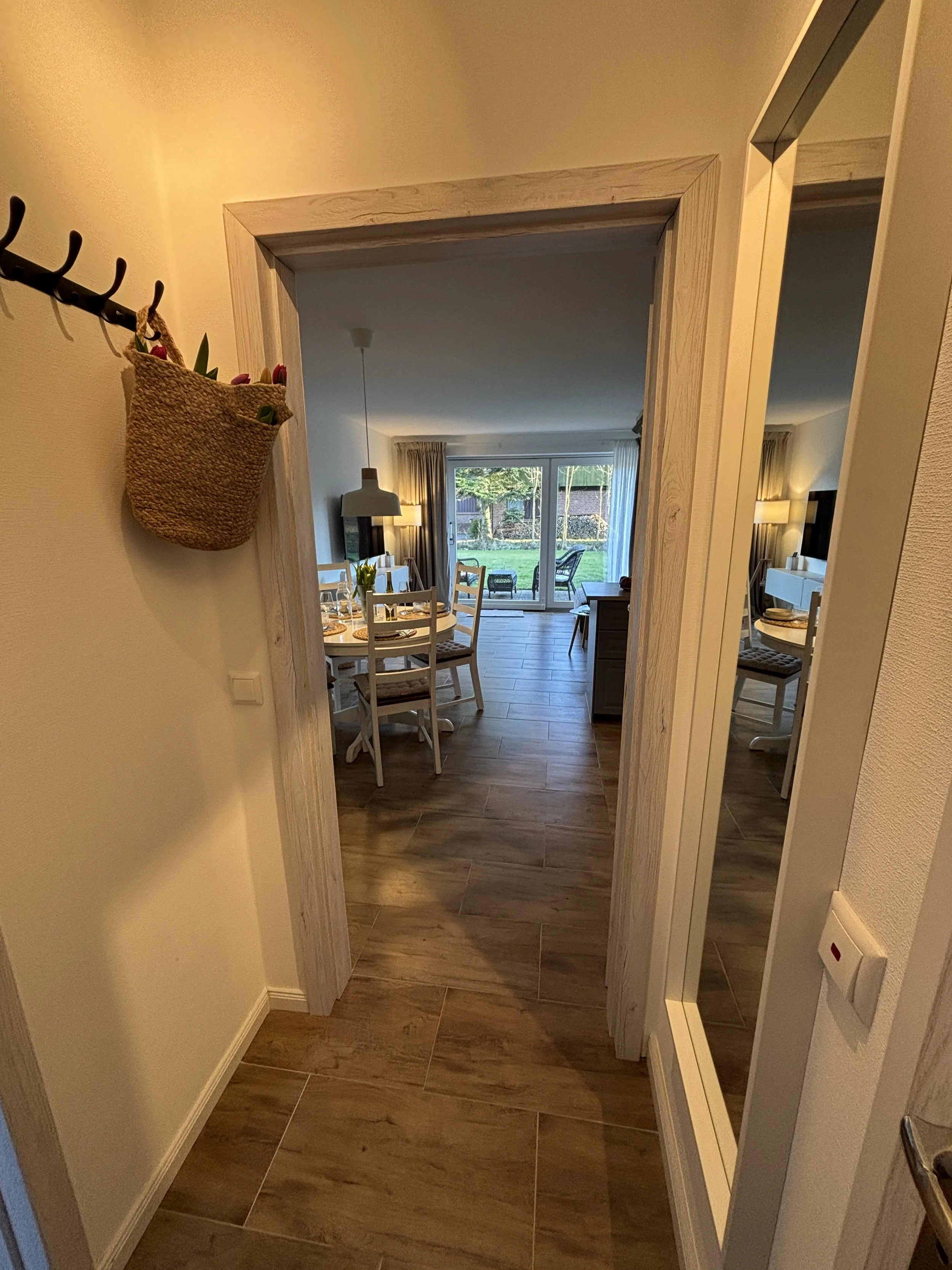 Ein Blick in ein Wohnzimmer mit Essbereich, große Fenster, Sicht auf den Garten, Holzfußboden, an der Wand befindet sich ein Haken mit einem Korb