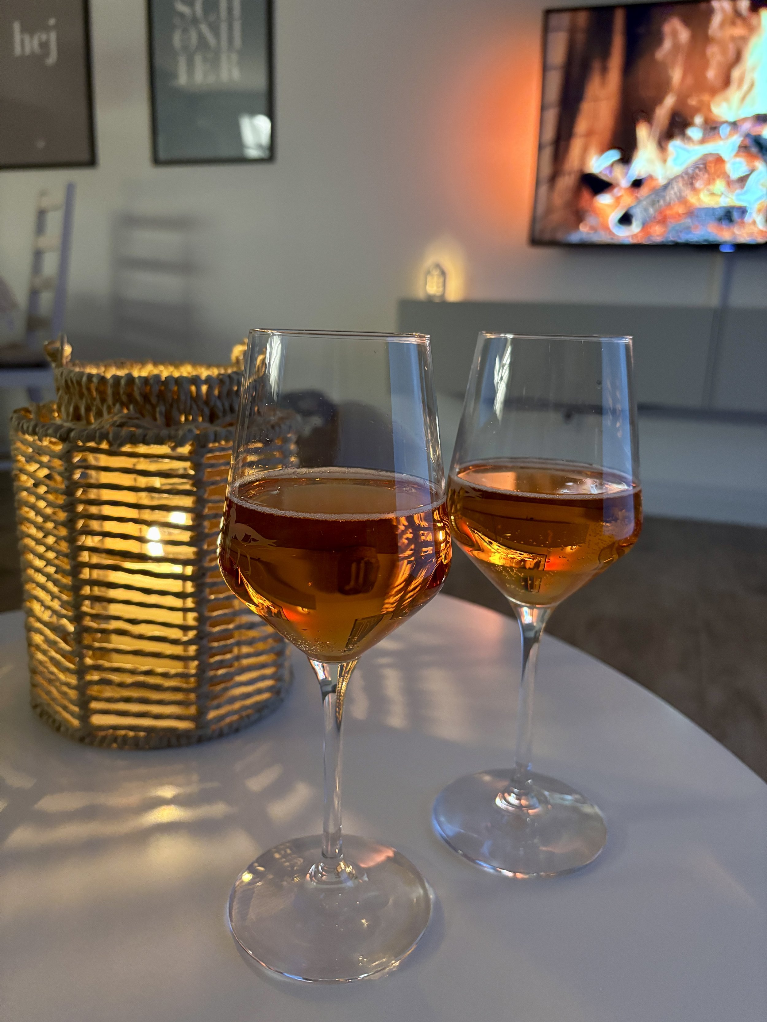 Zwei Gläser mit Roséwein auf einem weißen Tisch, im Hintergrund ein Korb mit Kerze und eine Fernseher, der ein Feuer im Kamin zeigt.