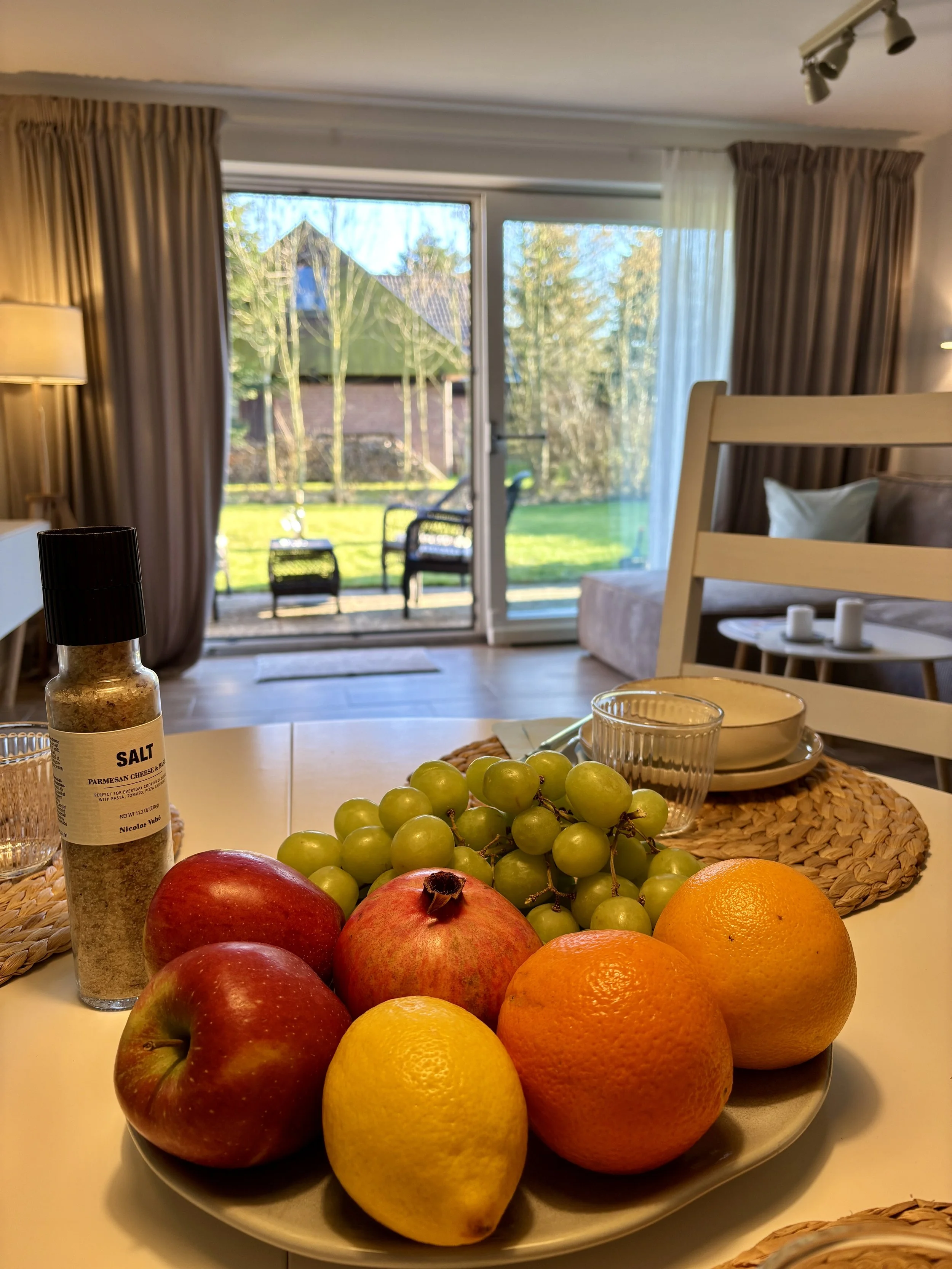 Früchte auf einem weißen Teller, darunter Äpfel, Orangen, Zitronen und ein Granatapfel, auf einem Esstisch im Wohnzimmer, neben einer Gewürzpackung und Trinkgläsern. Im Hintergrund ein offener Balkon mit Garten und Stühlen.