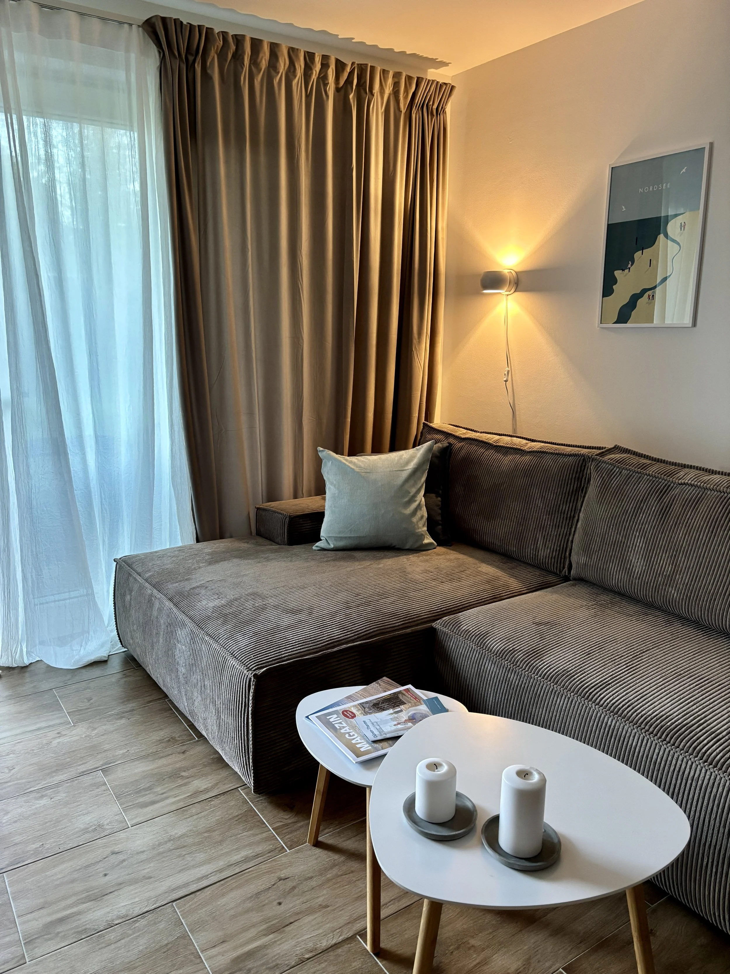 Ein gemütliches Wohnzimmer mit einem dunklen Sofa, einem kleinen weißen Couchtisch mit Kerzen und Magazinen, großen Vorhängen, einer Stehlampe an der Wand, und einem dekorativen Wandbild. Viel natürliches Licht durch die Fenster.