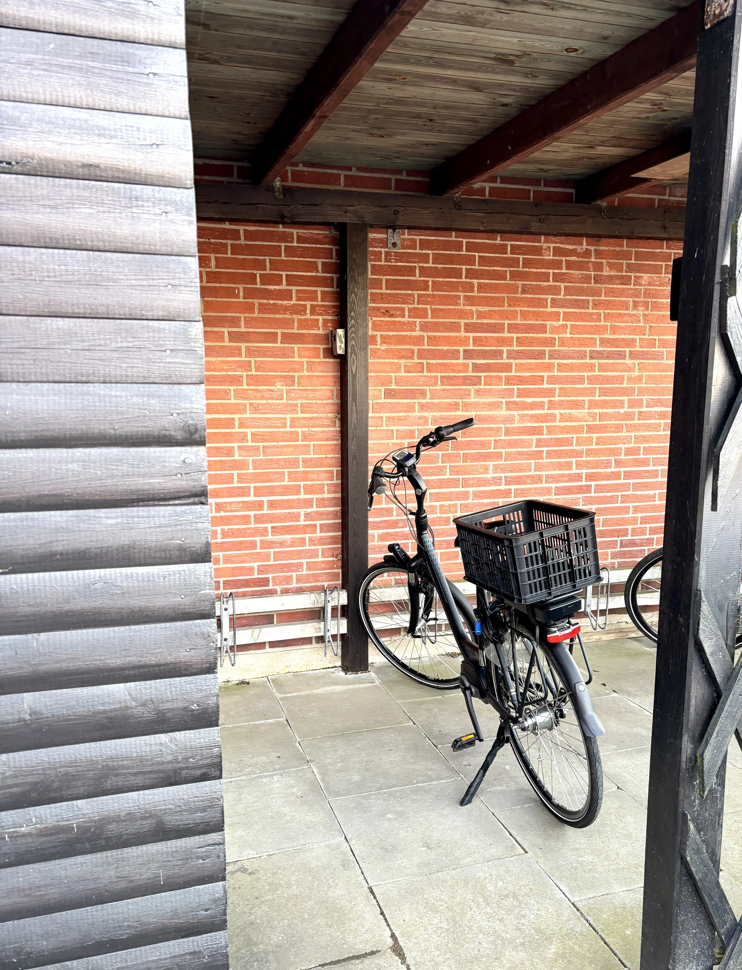 Ein Fahrrad mit Korb am Hinterrad, abgestellt auf einem gepflasterten Boden vor einer roten Ziegelwand, teilweise verdeckt durch eine Holzverkleidung auf der linken Seite.