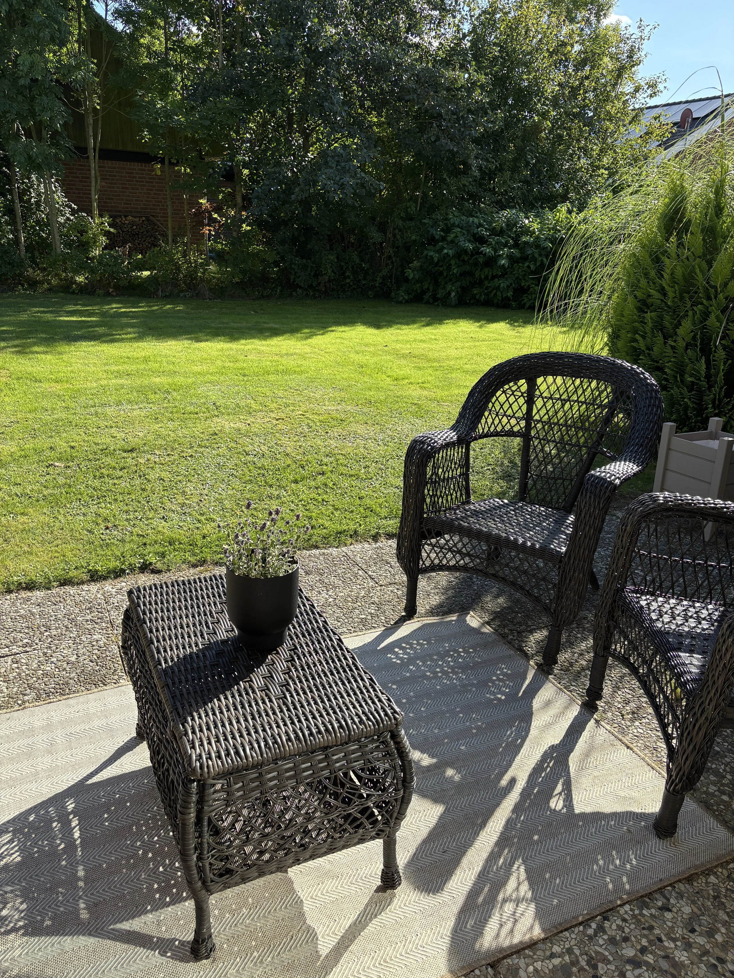 Garten mit grüner Rasenfläche, zwei schwarze Korbetten, ein kleiner Tisch mit einer topf mit Lavendel, im Hintergrund Bäume und Sträucher, sonniger Tag.