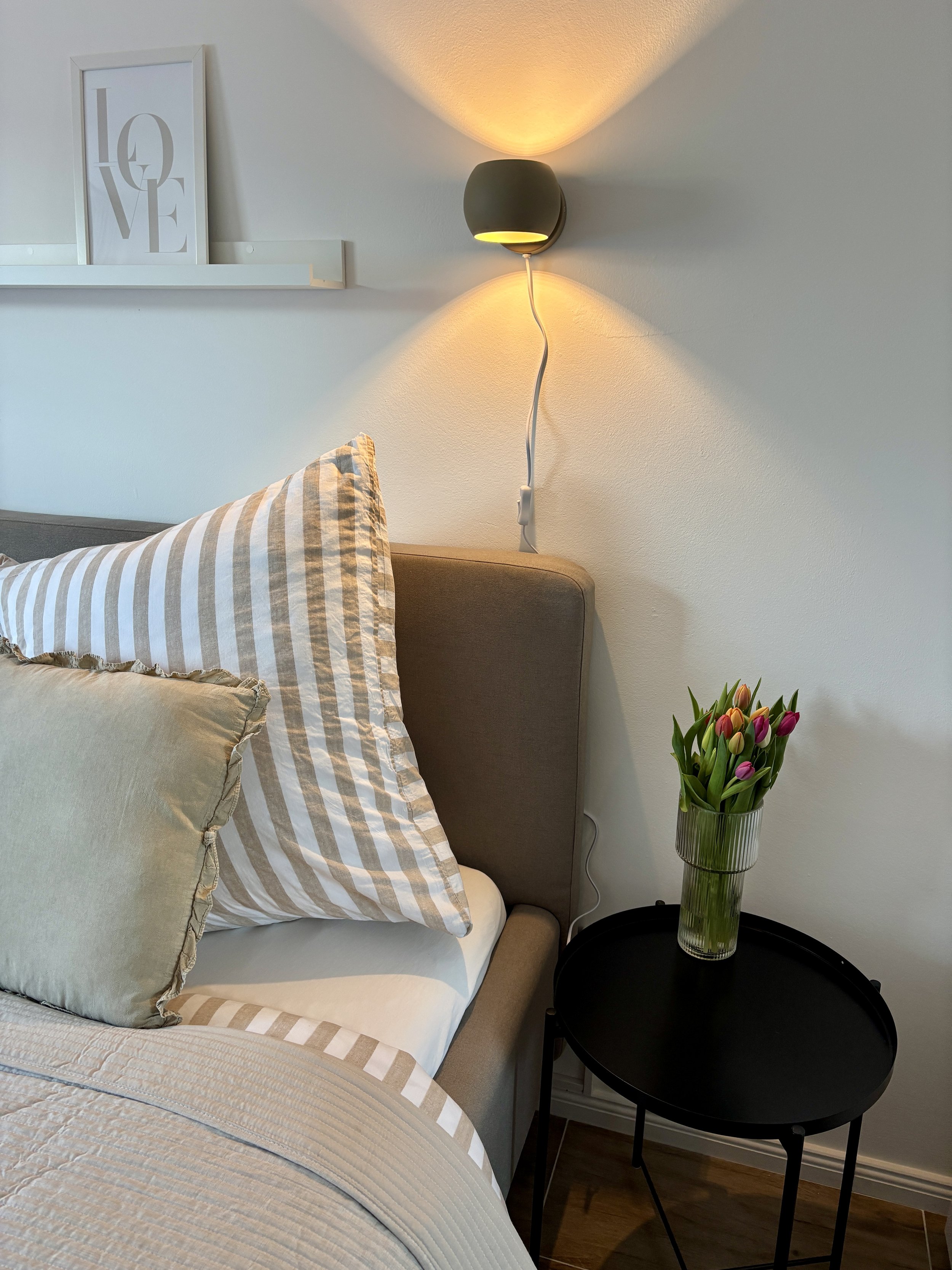 Ein Schlafzimmer mit einer Nachttischlampe an der Wand, einem Nachttisch mit einer Vase voller Tulpen, und einem Bett mit gestreiften Kissen.