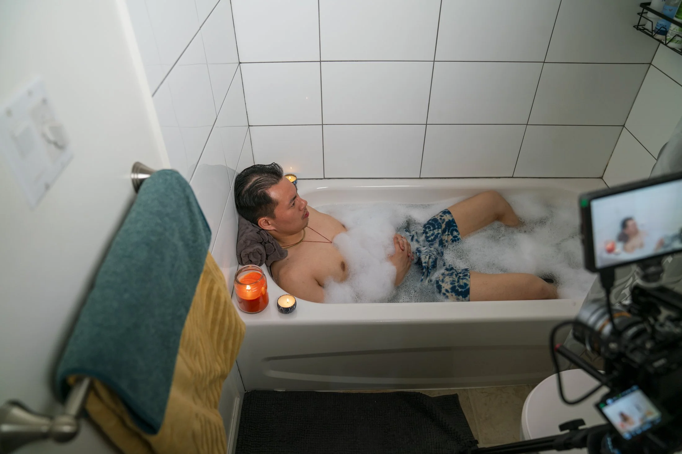 A man relaxing in a bubble bath with candles around the tub, being filmed in a bathroom. For a comedy scene