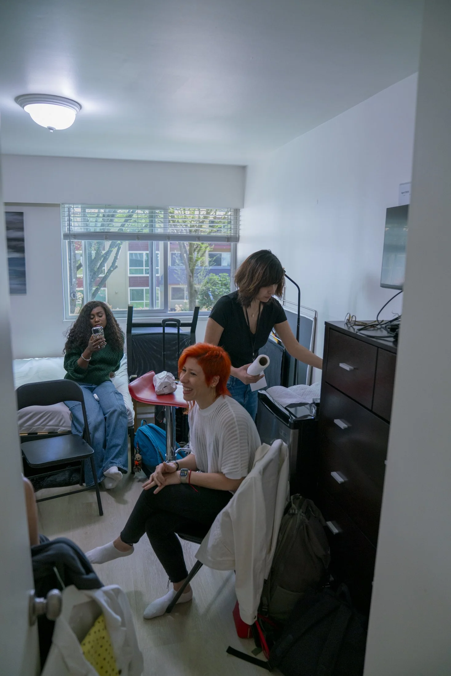 Three women in a small room with a window, surrounded by luggage and personal items, engaged in different activities. As they wait to go on a film set