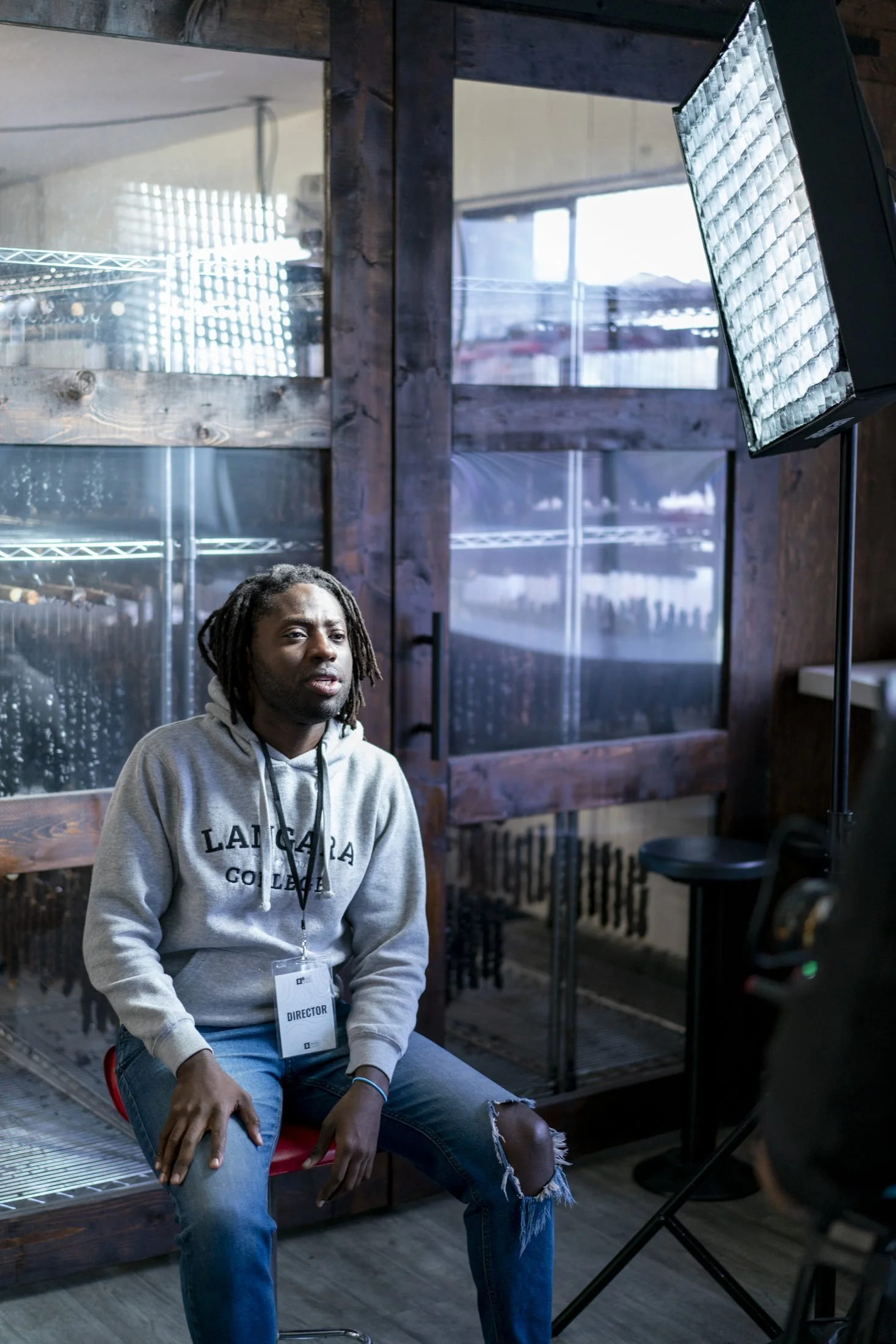 A man with dreadlocks wearing a gray hoodie and ripped jeans sitting on a stool during a video shoot or interview, with professional lighting and equipment in a rustic indoor setting.