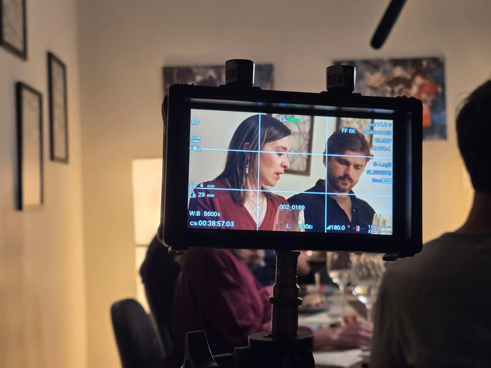 A behind-the-scenes view of film directors during a shoot, with camera monitor showing a woman with shoulder-length dark hair and dangling earrings, and a man looking over her shoulder at a dinner table.
