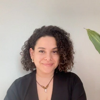 A woman with curly hair smiling at the camera, wearing a black top, jewelry including earrings and a necklace, with a potted plant to her right.