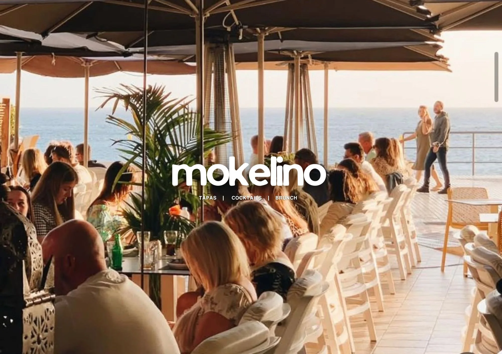 Personas en una terraza con vista al mar disfrutando en un restaurante abierto, con sombrillas y decoración tropical.