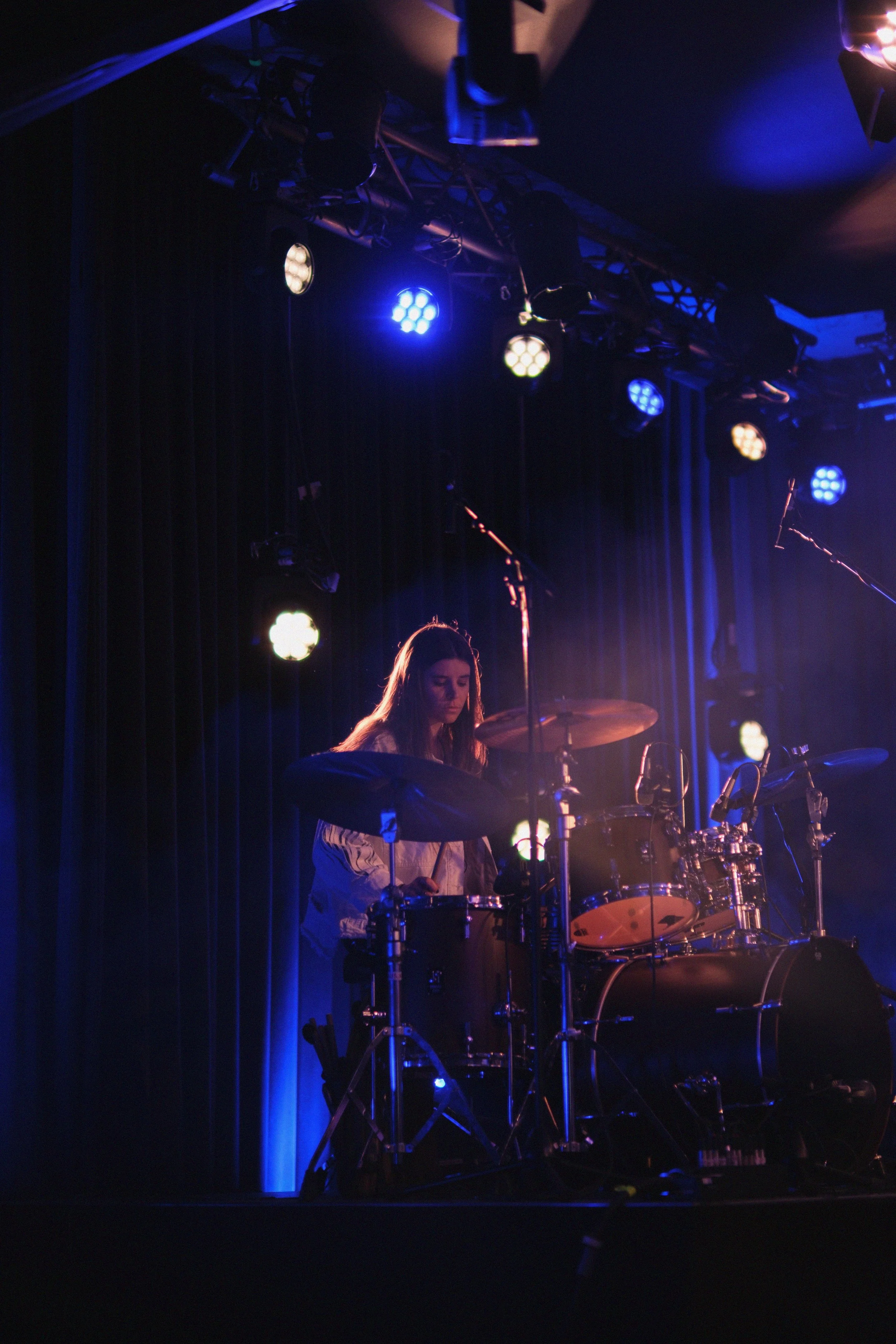 A female drummer playing a drum set on stage, illuminated by blue and purple stage lights, with a dark background and stage equipment overhead.
