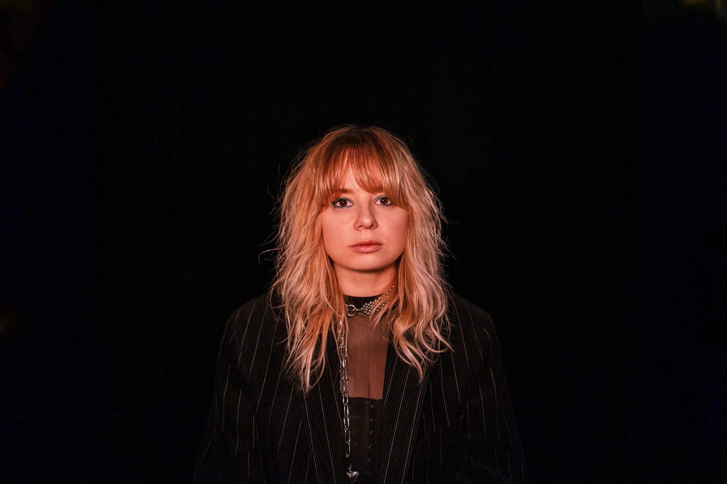 A young woman with blonde, wavy hair wearing a black pinstripe blazer and a chain necklace, standing against a black background.