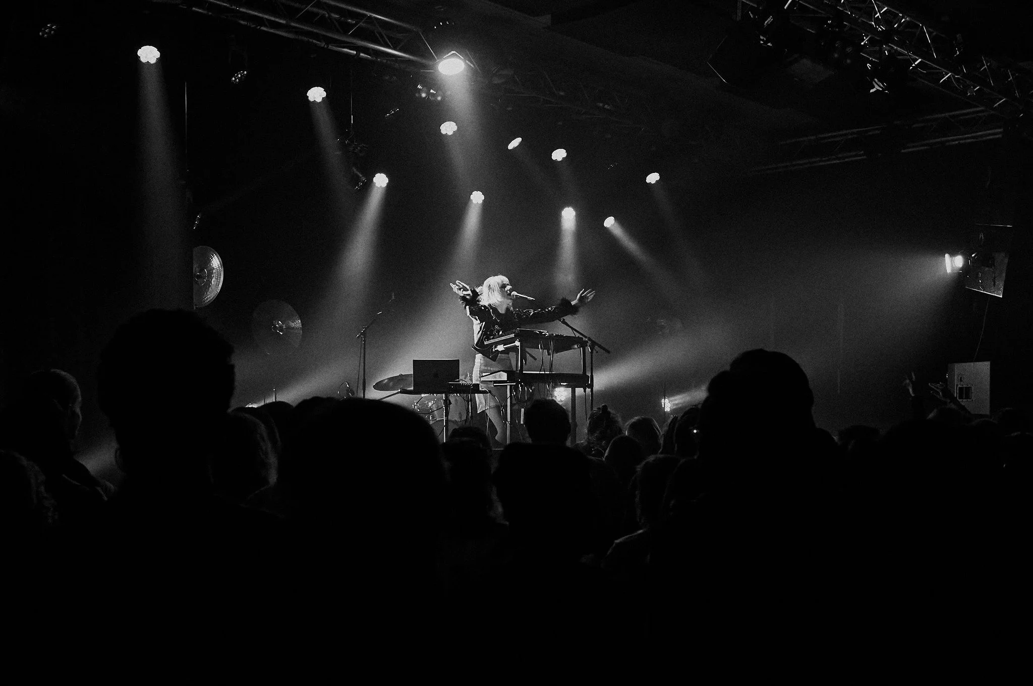 A black and white photo of a live music performance on stage featuring a female singer with long hair, performing at a keyboard and microphone. Audience silhouettes are visible in the foreground, and stage lighting is focused on her.