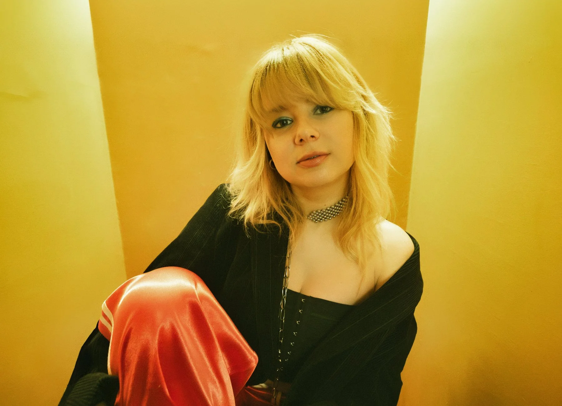 A woman with blonde hair and blue eyes wearing a black off-shoulder top, a pearl choker, and holding a red satin piece of clothing, sitting against a yellow wall.