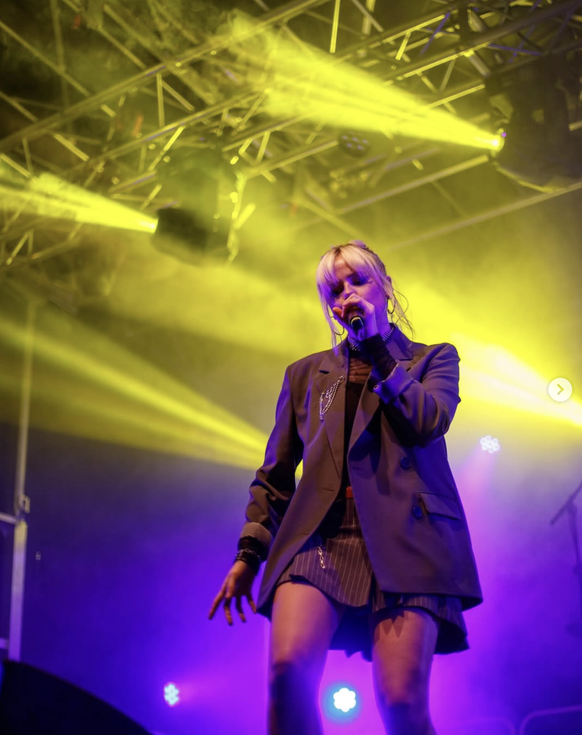 A female singer performing on stage under yellow and purple stage lights, holding a microphone, wearing a blazer and striped shorts.