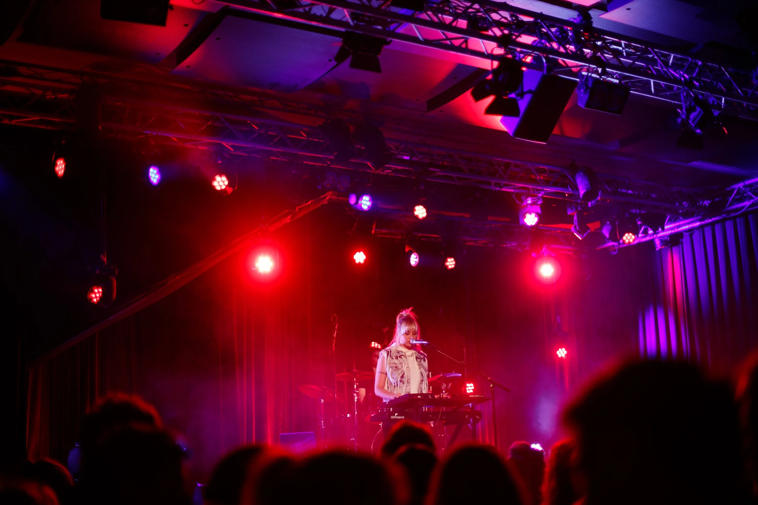 A female musician performing on stage at a concert, illuminated by red and purple stage lights, with an audience watching.