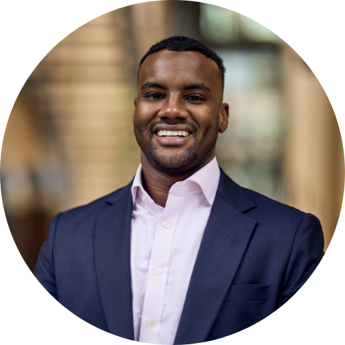 A smiling man in a dark blue suit and white shirt standing outdoors in an urban setting.