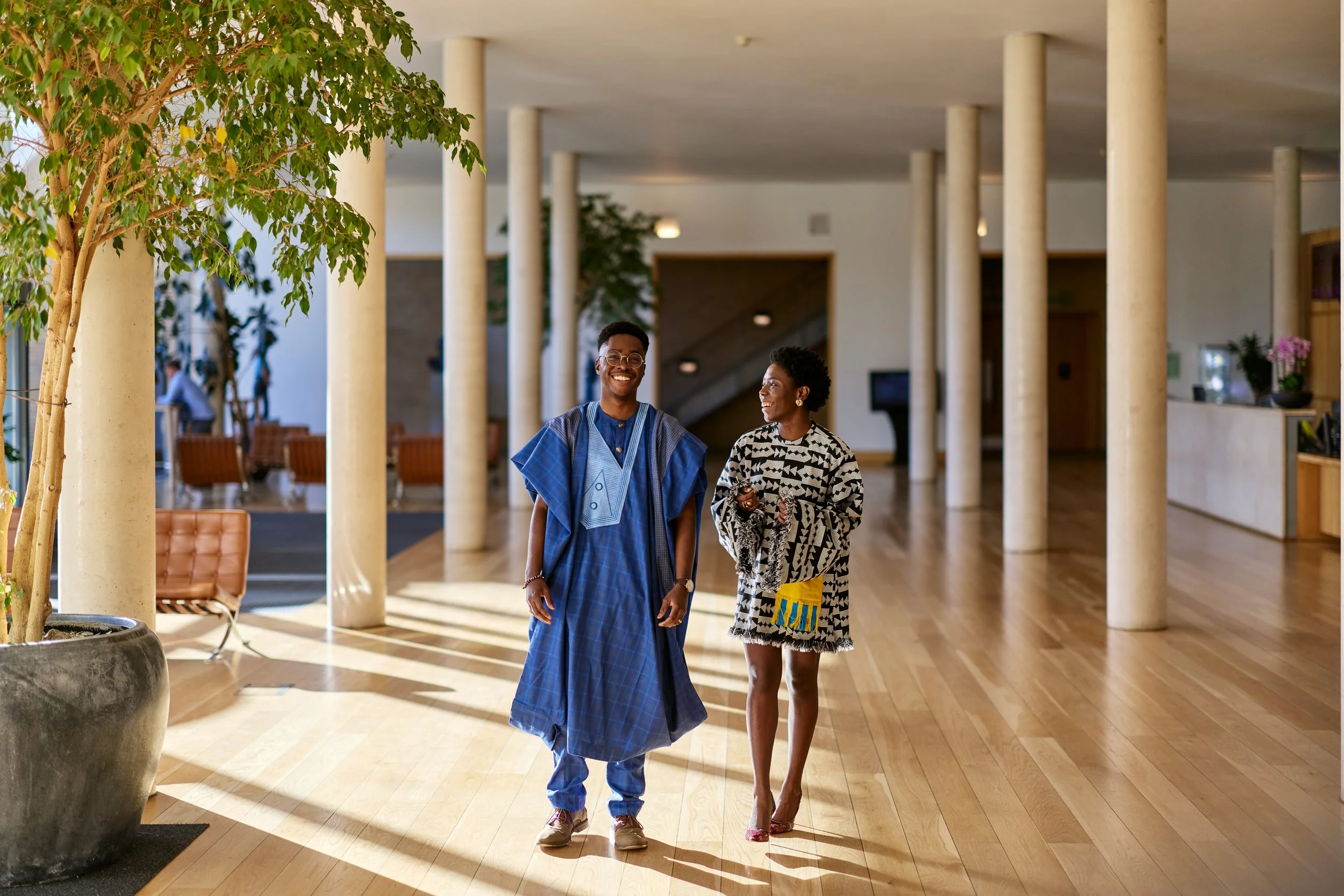 Two members of Oxford Africa Business Alliance smiling and walking in a spacious, well-lit lobby or reception area with wooden floors, large potted plants, and modern decor.