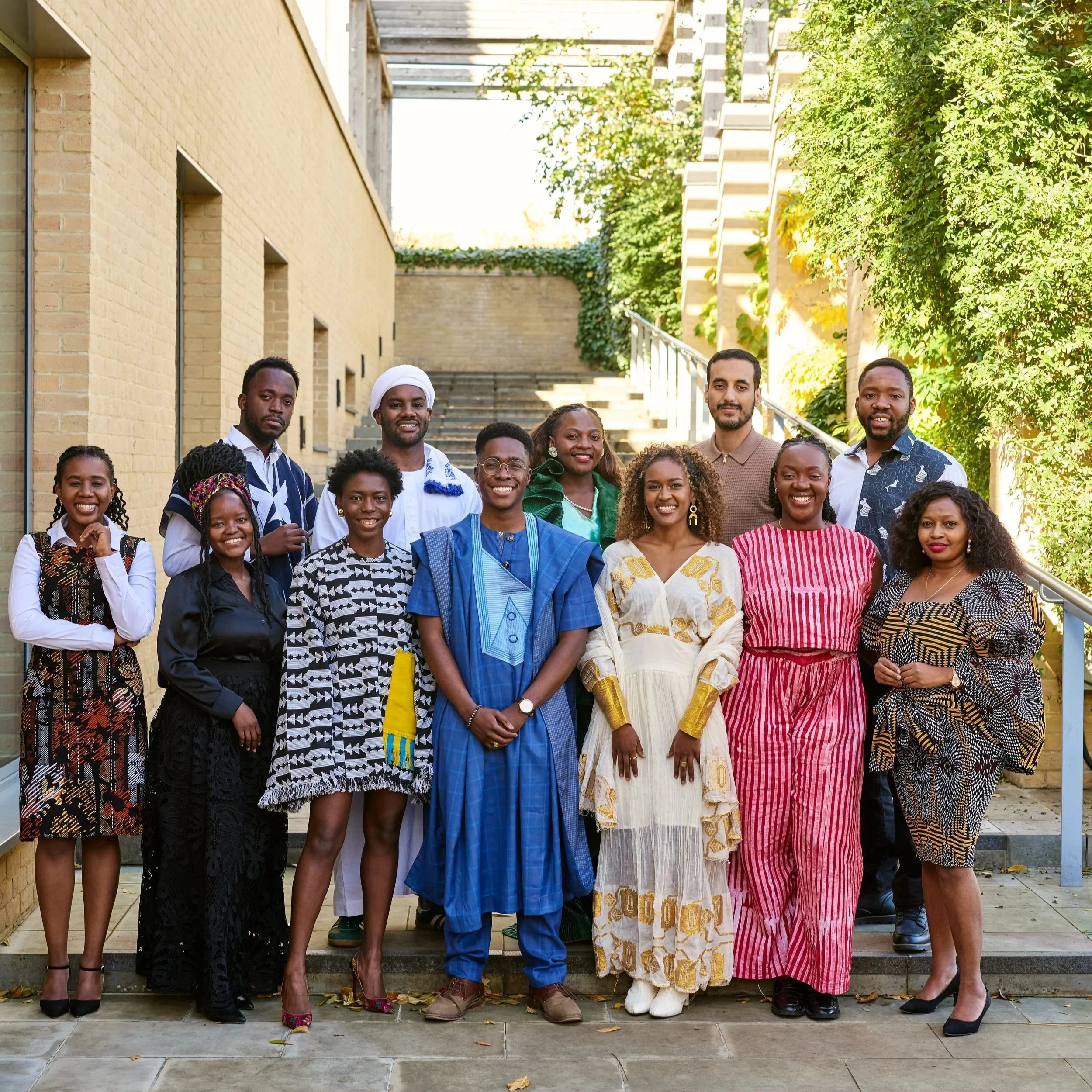 Group of twelve diverse people standing outdoors on steps with greenery and brick walls in the background, smiling at the camera.