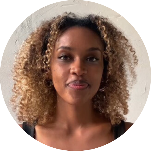 A woman with curly blonde hair, wearing hoop earrings and a black top, standing against a white wall.