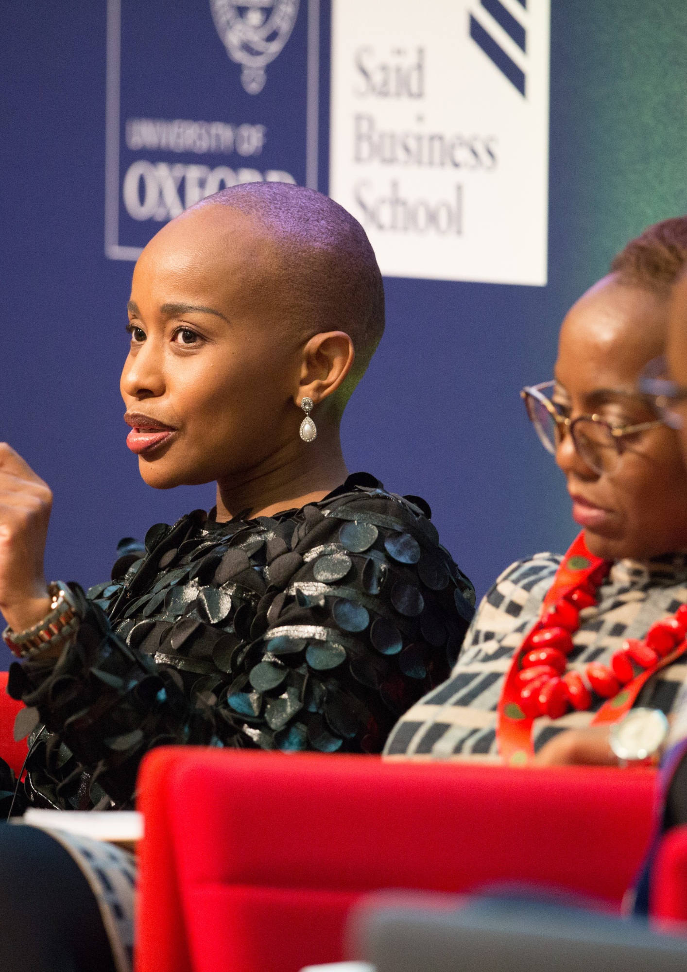 Two women seated at a panel or conference, with a blue background featuring a sign that reads 'University of Oxford' and 'Sand Business School.' One woman is wearing a black, sequined top and large earrings, while the other is wearing glasses, a patterned outfit, and a red necklace.